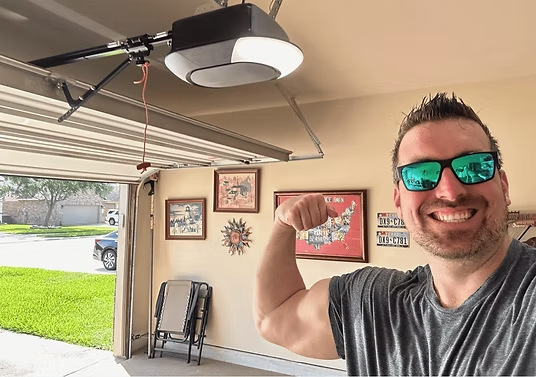 A smiling man wearing sunglasses flexing his arm in a garage with a garage door opener, framed pictures, and a lawn visible outside.
