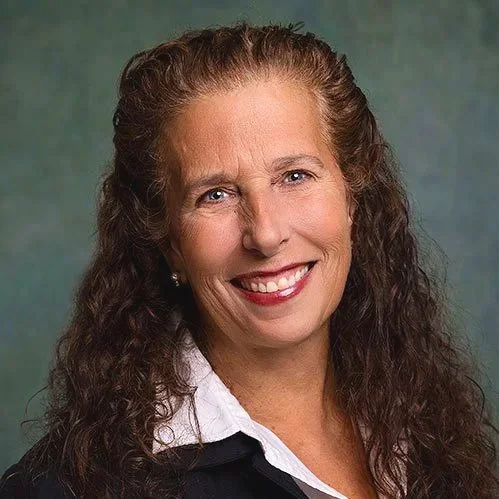 A woman with long, curly brown hair smiling and wearing a white collared shirt and black blazer.