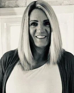A woman with straight blonde hair, smiling, wearing a dark cardigan over a light top, in front of a kitchen background.