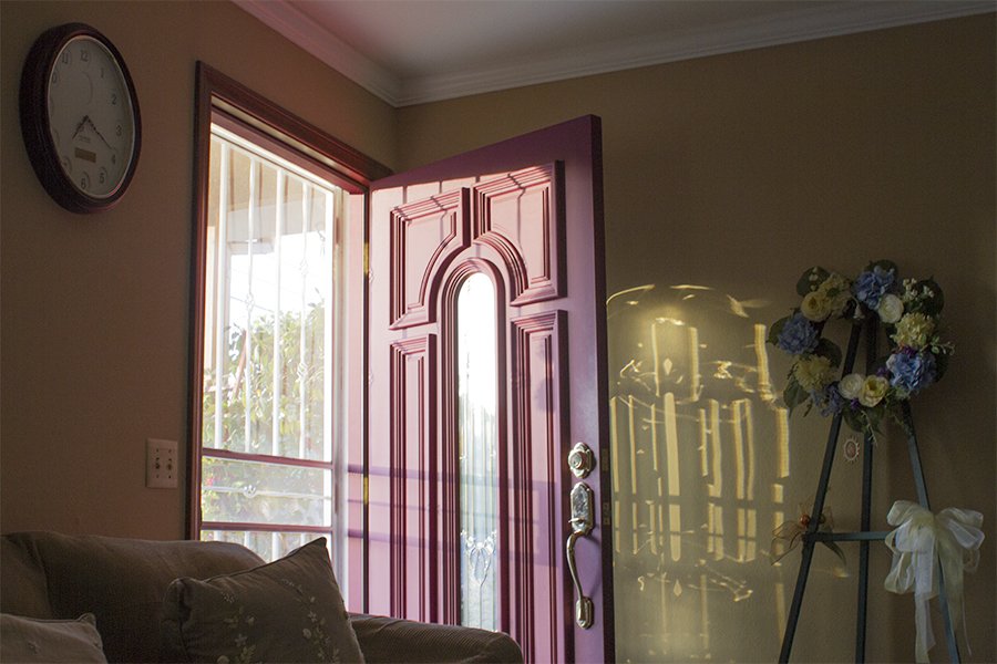 Open pink front door with sunlight casting shadows inside a living room. A round wall clock, a sofa with pillows, and a flower wreath on a stand sit inside.