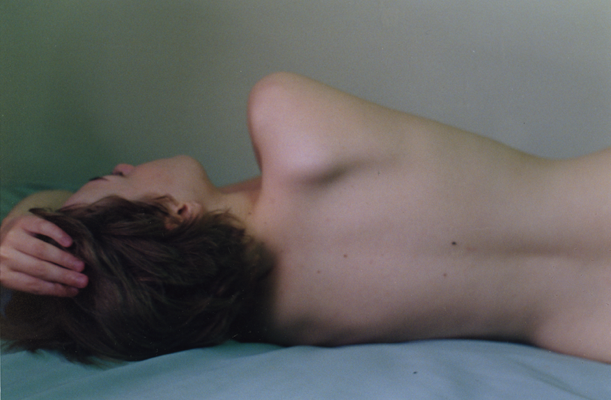 A topless woman with brown hair lying on her side on a bed, with one hand on her head and her eyes closed.