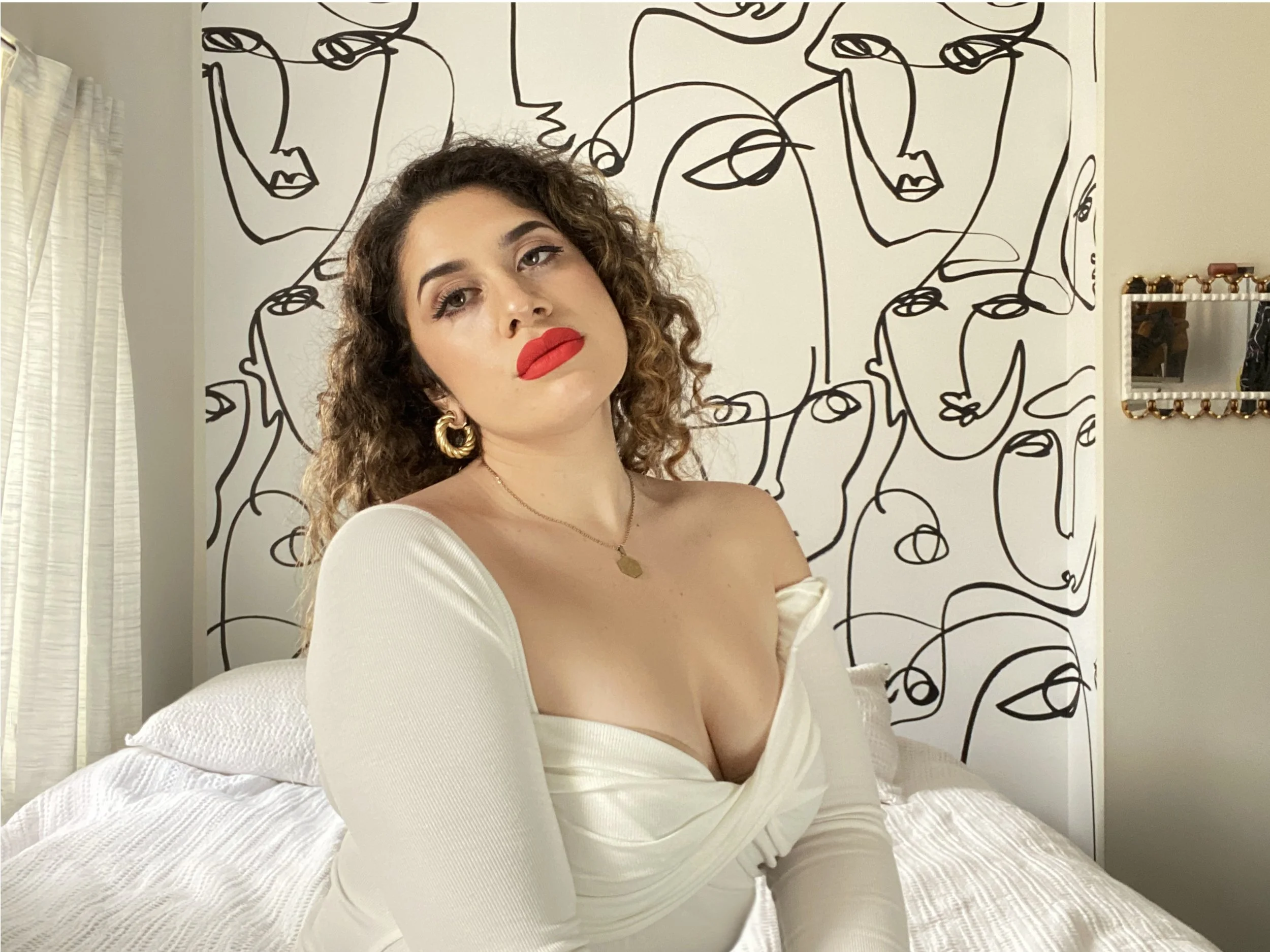 A woman with curly hair and red lipstick sitting on a bed in a white off-shoulder dress, with a black line abstract face art mural in the background.