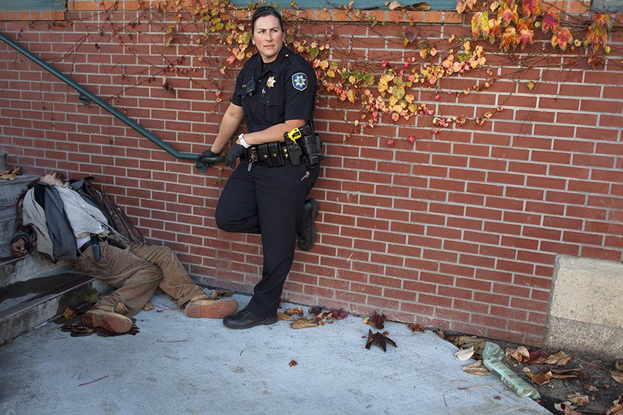 A police officer standing against a brick wall with autumn leaves, holding a stick, next to a homeless person lying on the ground.