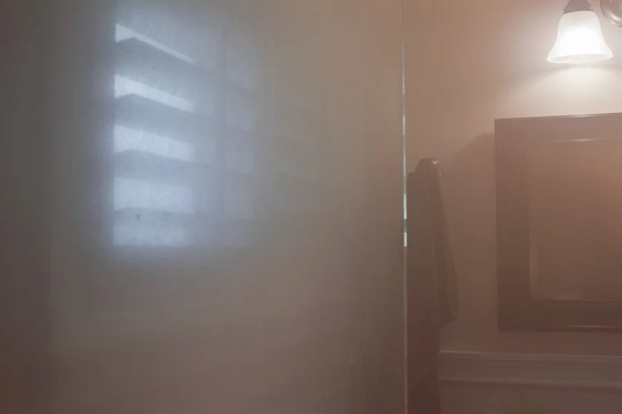 A reflection of a window with blinds on a mirror or shiny surface in a dimly lit room, with a wall-mounted light fixture and a television visible in the background.