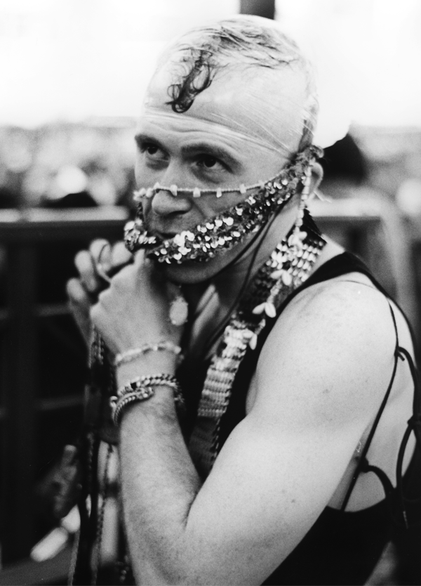 A person with a bald head and a curl of hair on top, wearing a garland and a beaded necklace around the neck, with extra beads and jewelry on the hands and wrists. They are seen in black and white, with a supporting strap on the left shoulder, and ha