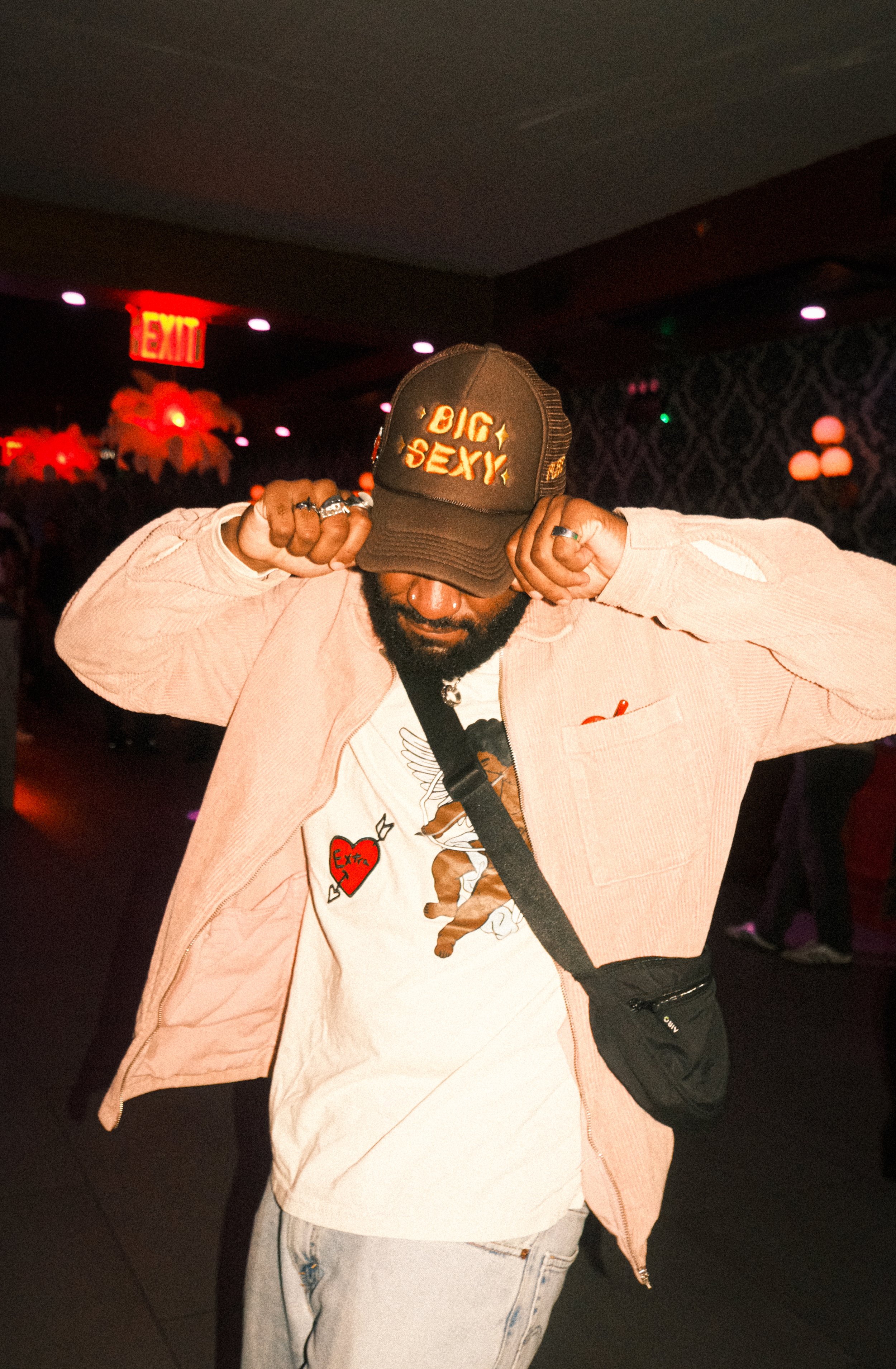 A man with a beard adjusting his cap, wearing rings and a pink jacket, in a dimly lit room with red and purple lighting, and a neon exit sign in the background.