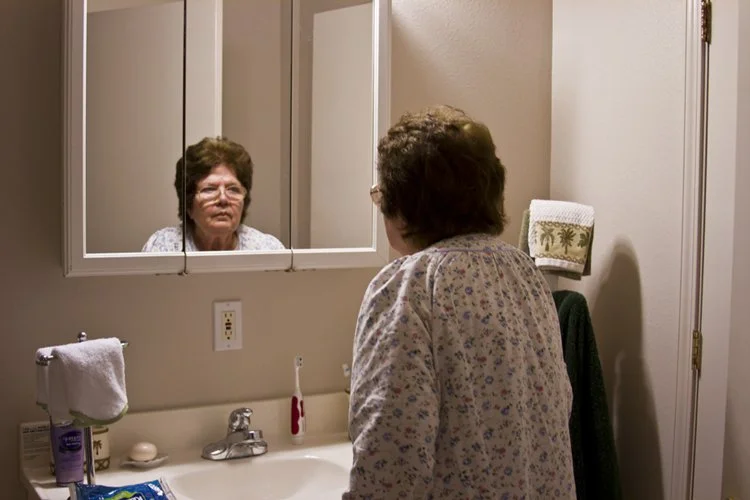 An elderly woman standing in front of a mirror in a bathroom, looking at her reflection.