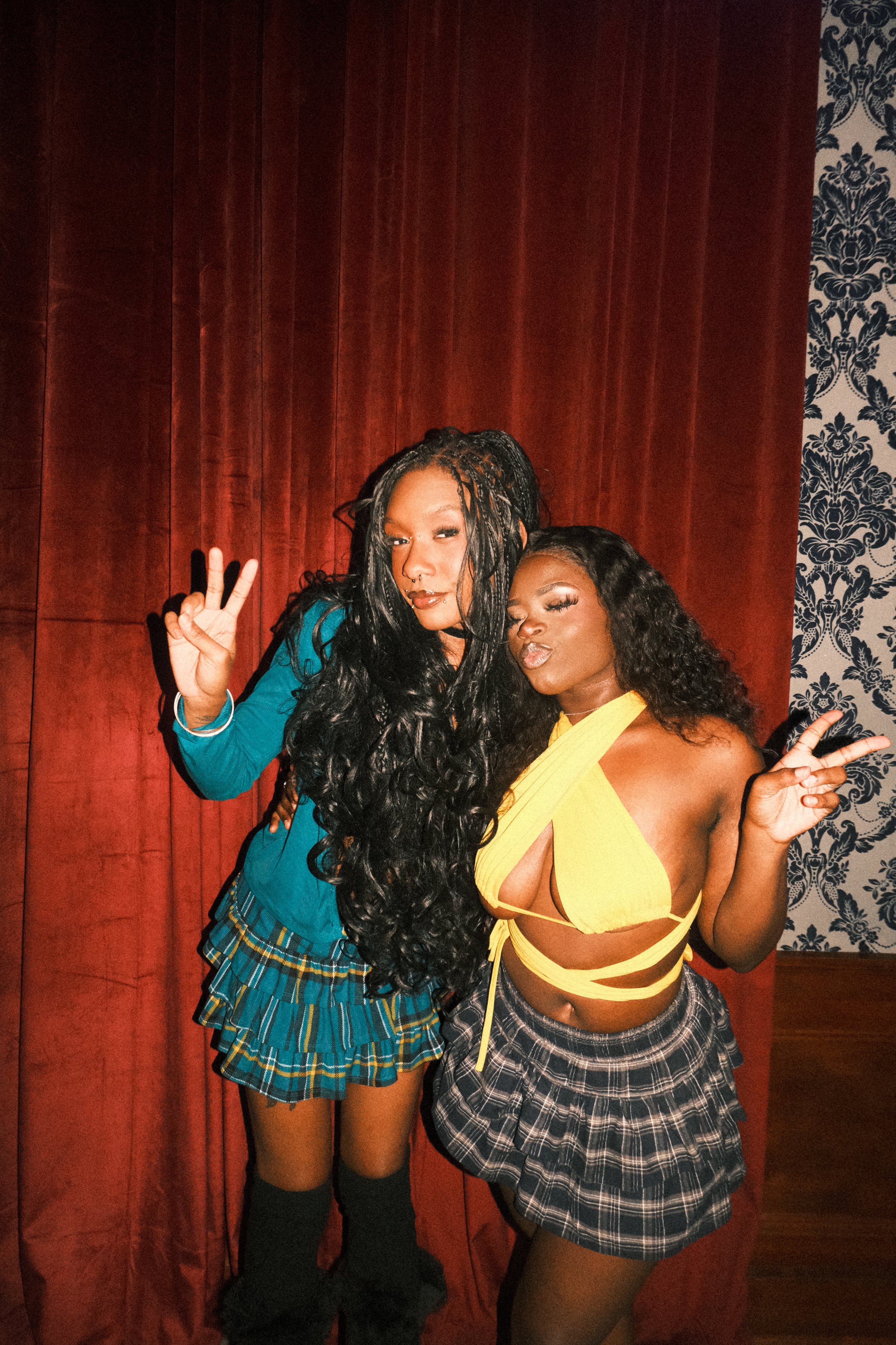 Two women posing together, making peace signs with their hands, standing in front of a red curtain and patterned wallpaper.
