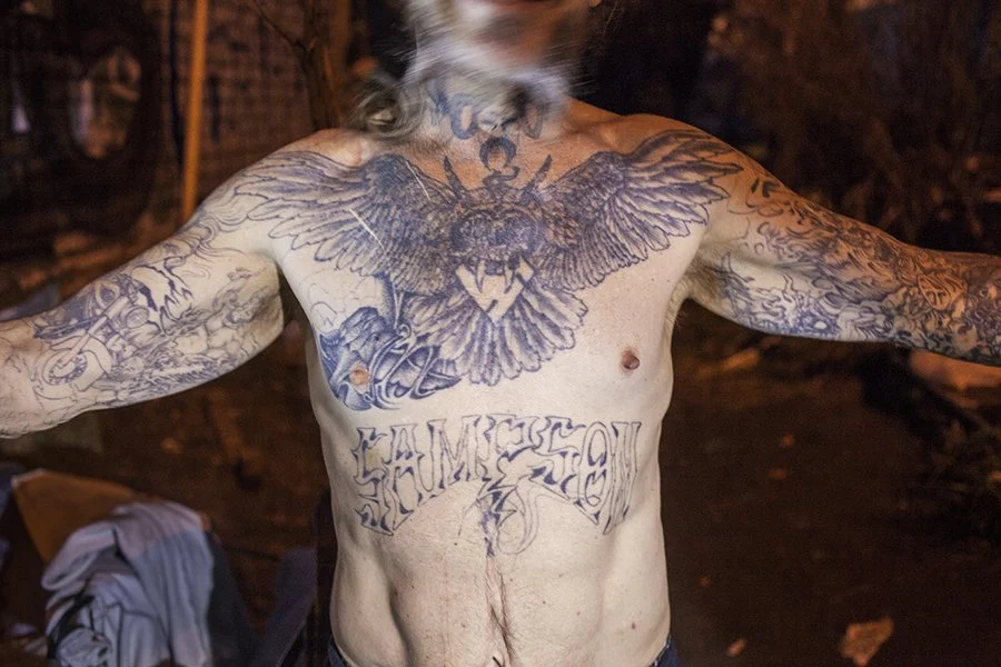 A shirtless person with tattoos on their chest and arms, including a large eagle with outstretched wings on the chest and the words "HUMBLE SO" below it. The person is standing with arms outstretched in a dimly lit setting.