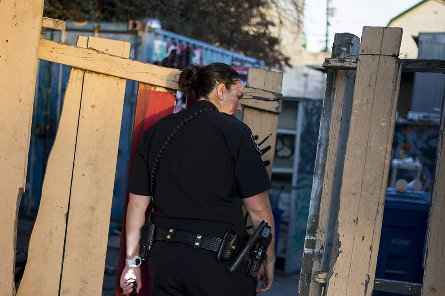 A police officer walking past a makeshift wooden fence in an outdoor setting, possibly an alley or yard.
