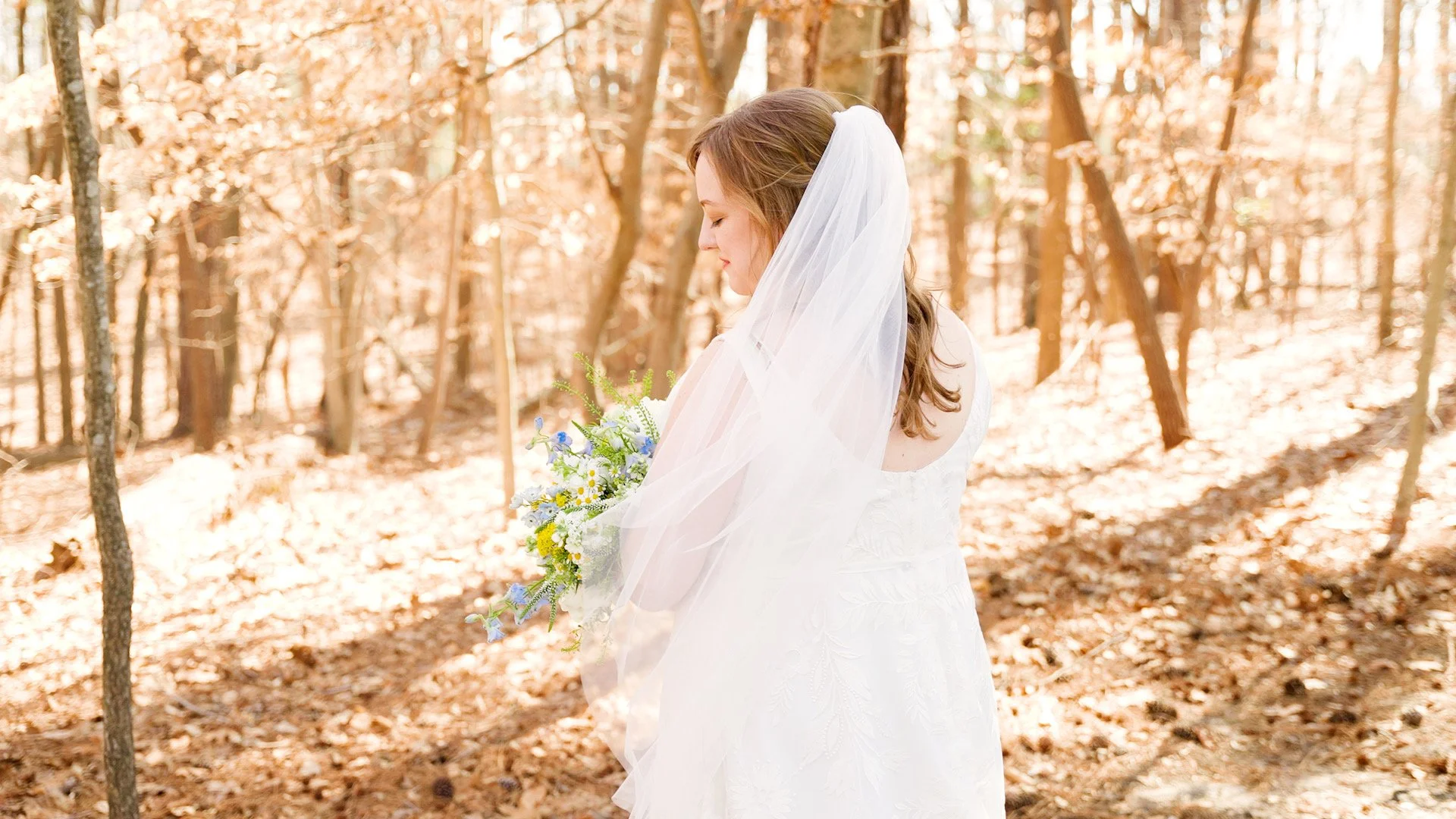 A bride stands in a sunlit forest holding a bouquet of flowers, with her eyes closed and wearing a white wedding dress and veil.