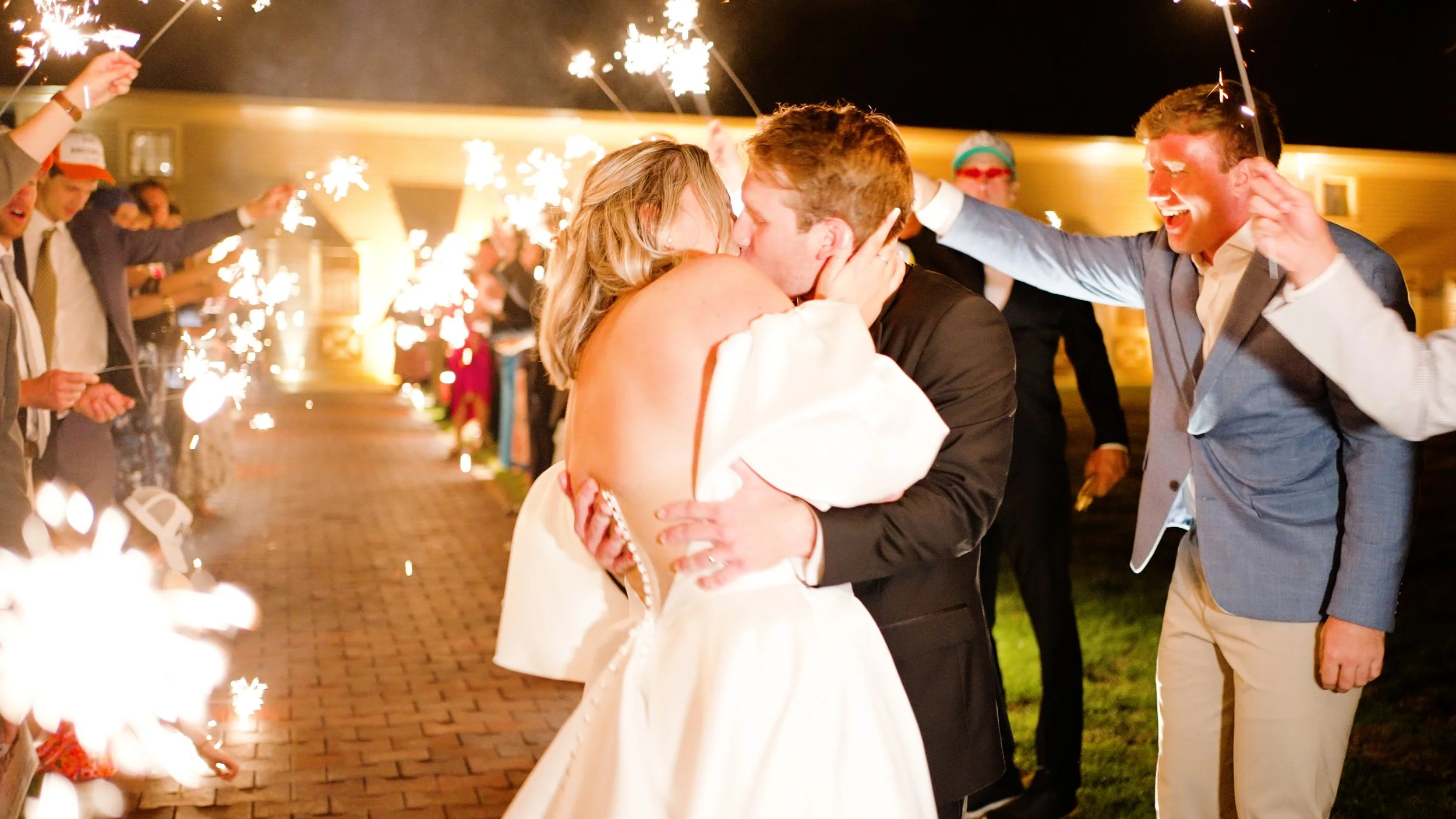 A newlywed couple shares a kiss while being surrounded by friends and family holding sparklers during a nighttime celebration.
