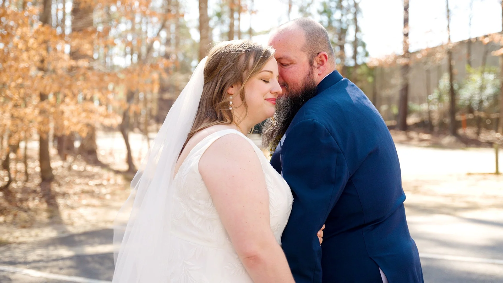 A newly married couple sharing an intimate moment outdoors, with eyes closed and smiling gently, surrounded by autumn trees.
