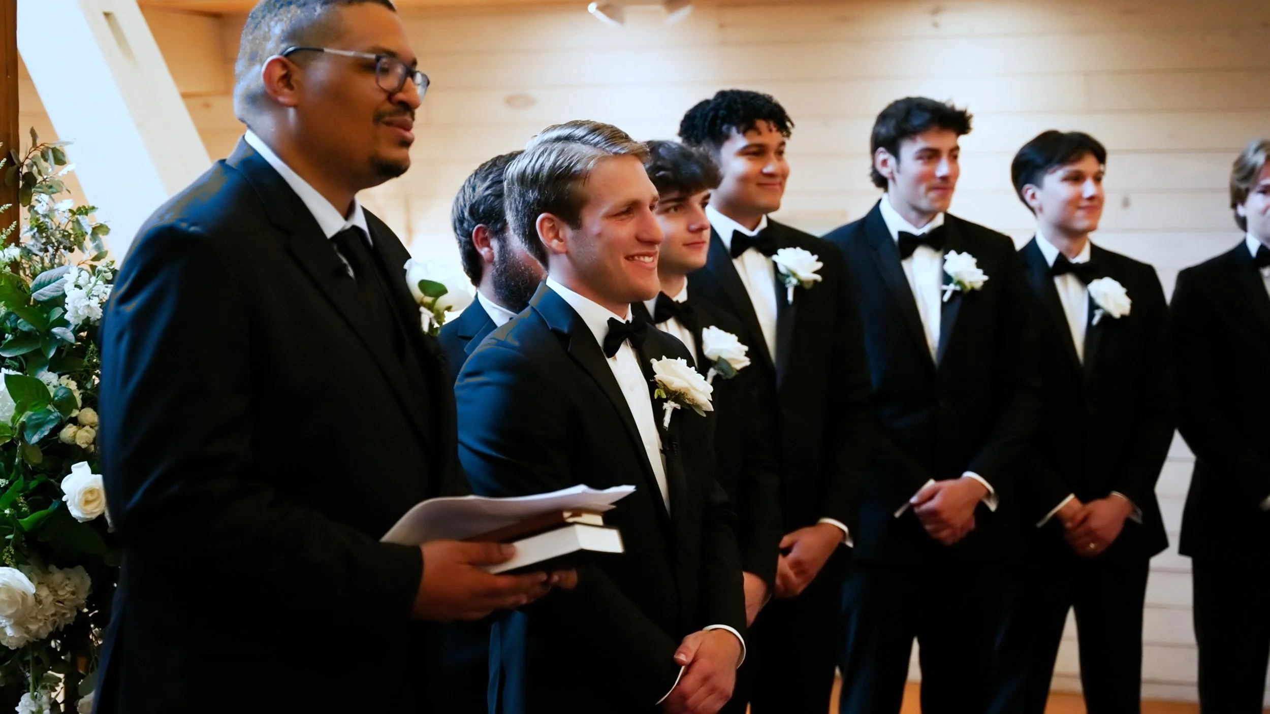 Groom and groomsmen dressed in black tuxedos with white shirts and black bow ties, standing in a row during a wedding ceremony.