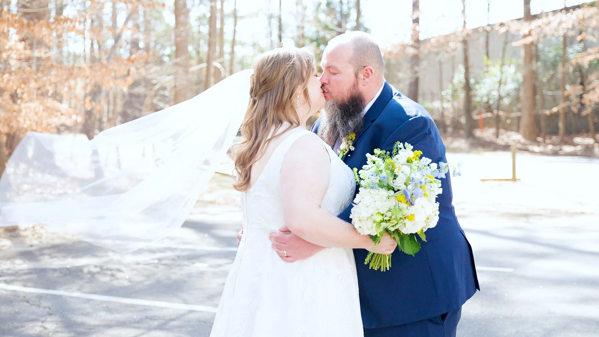 A bride and groom share a kiss outdoors on their wedding day, with the bride holding a bouquet and a veil flowing in the wind.