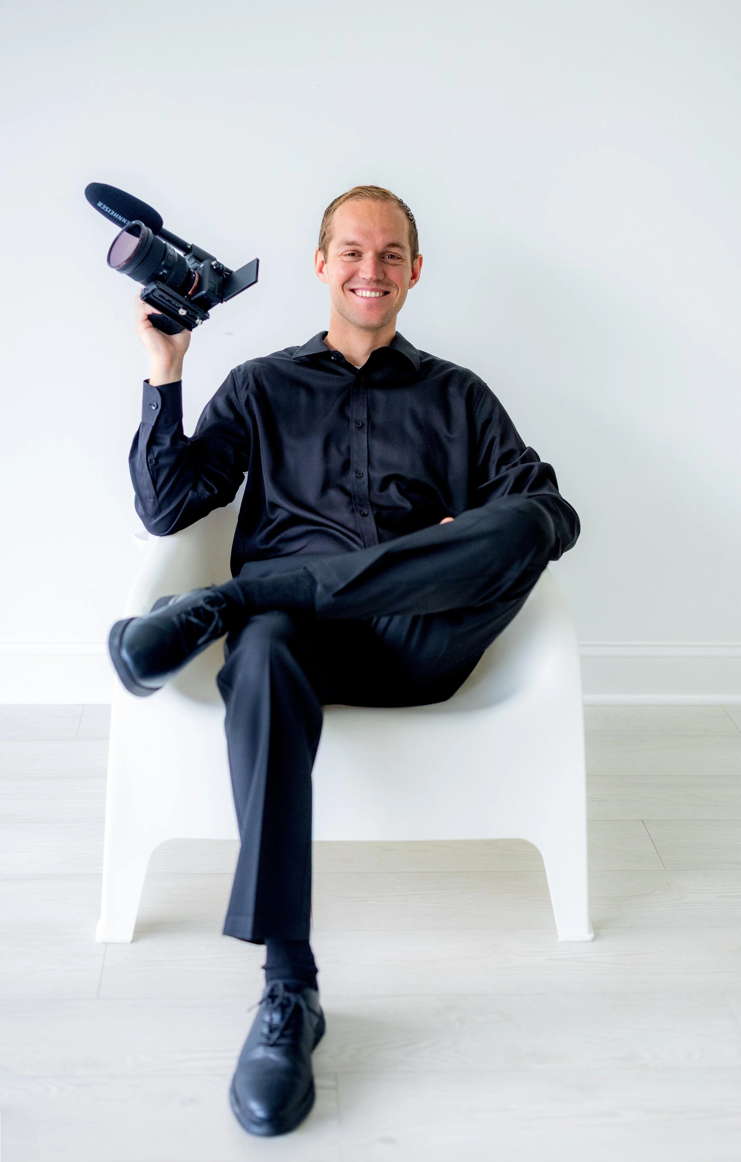A man in a black shirt and black pants sitting cross-legged on a white chair, smiling, holding a camera with a microphone attached, against a plain white background.