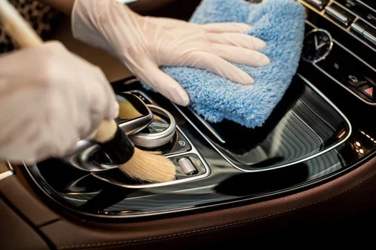 Person cleaning the center console of a car with a microfiber cloth and a brush while wearing gloves.