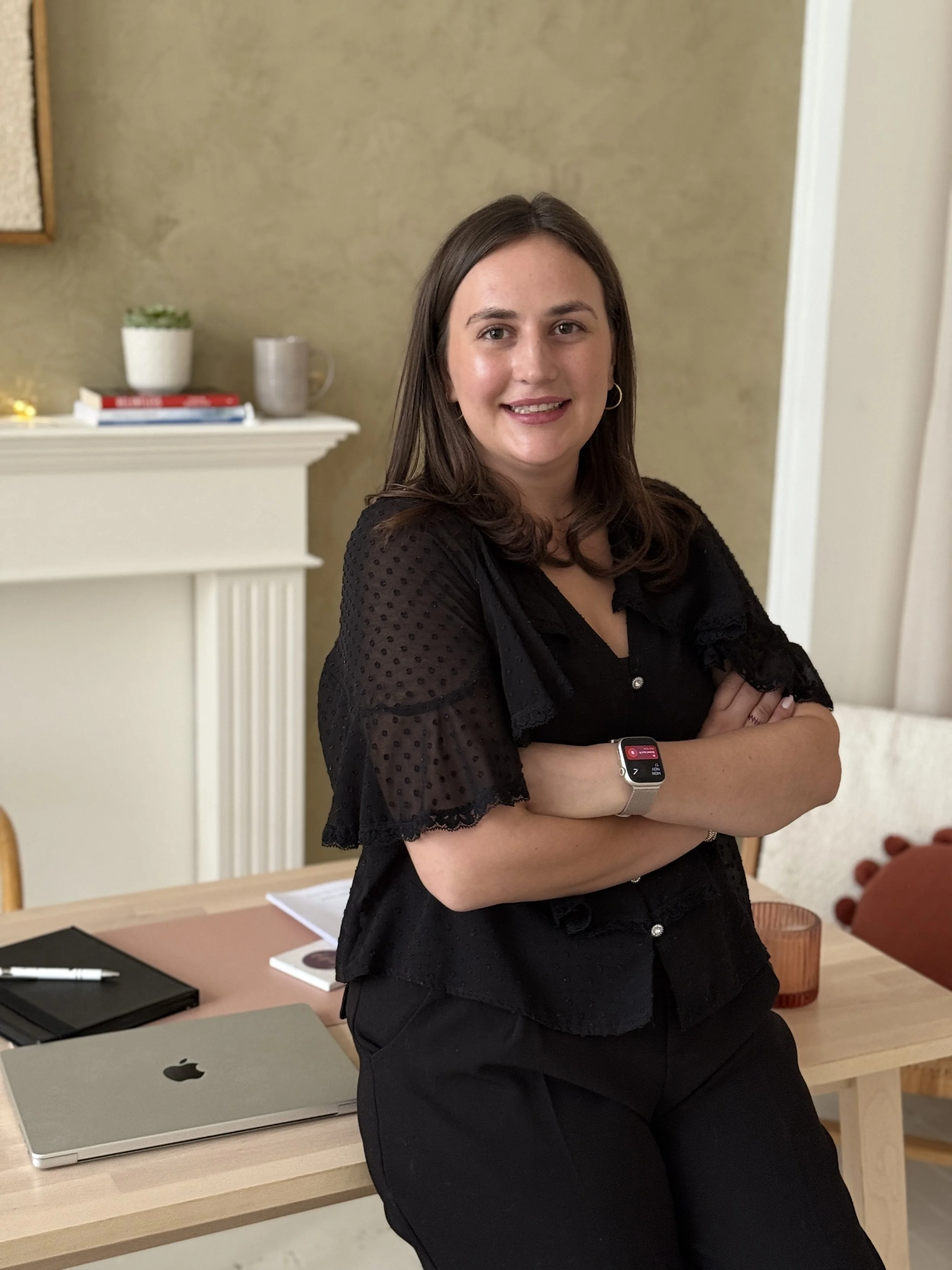 A woman with shoulder-length brown hair, wearing a black blouse with sheer, polka-dotted sleeves, standing with crossed arms in a room with a beige wall, a white fireplace mantel with books, a small potted plant, and a grey mug, and a wooden table with a closed MacBook, black notebooks, and a pen.