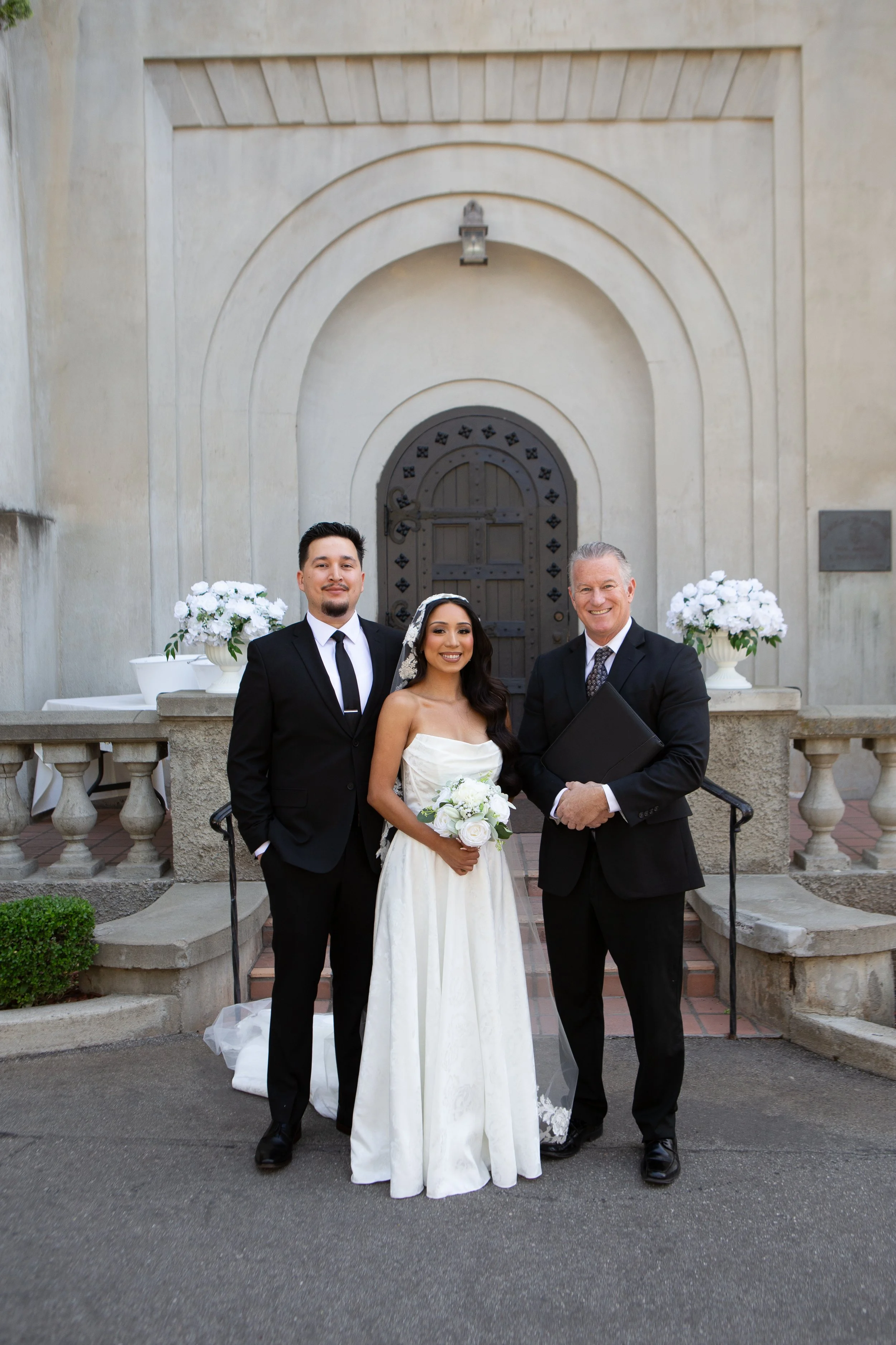 A wedding scene with three people, a bride in a white wedding dress holding a bouquet, and two men in suits, standing in front of a historic building with an arched wooden door and decorative stonework, floral arrangements on either side.