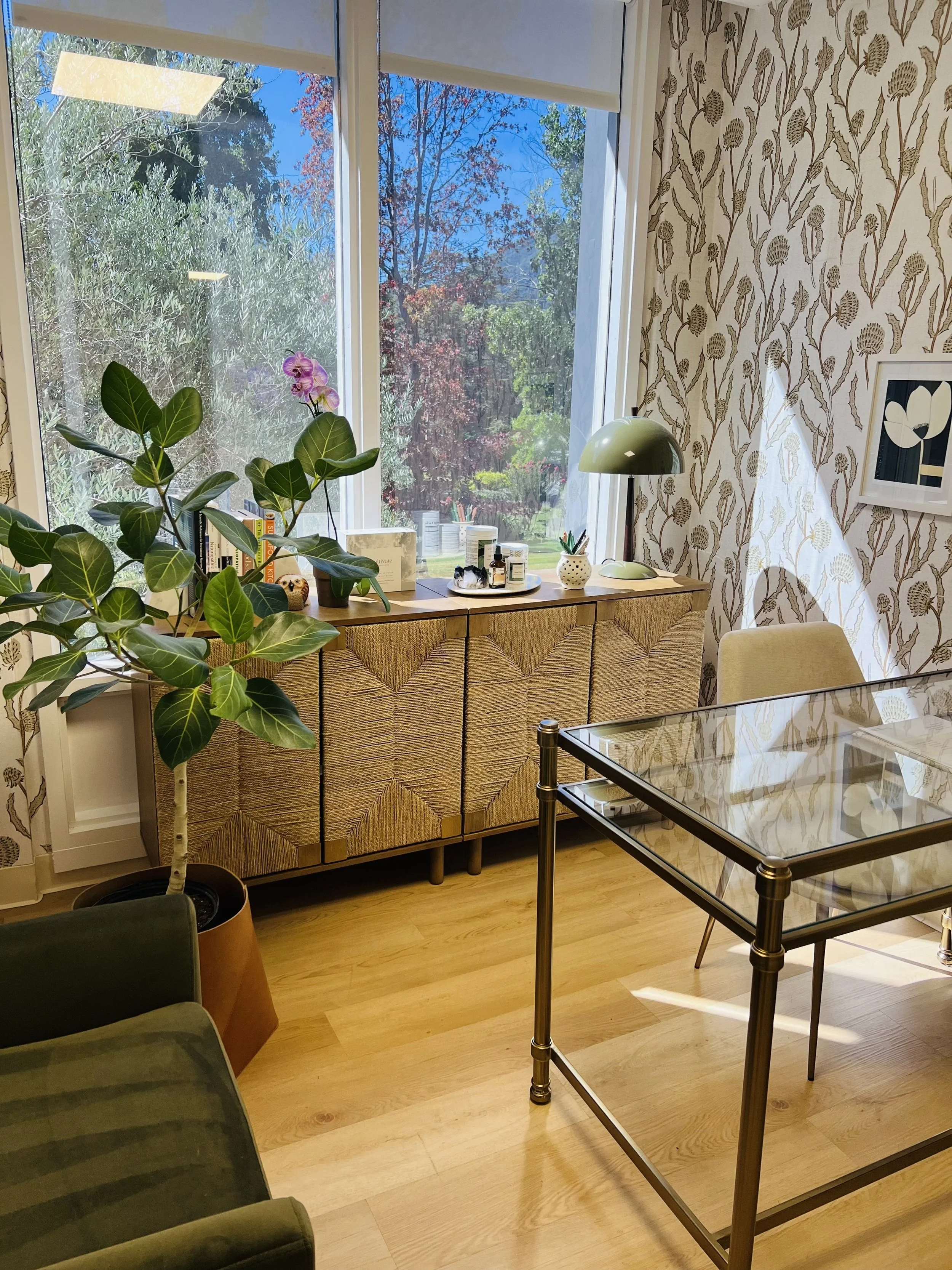 A room with a large window showing trees outside, a wooden sideboard with decorative items and essentials on top, a potted plant, a glass-top table, and a cushioned chair. The walls have floral-patterned wallpaper, and sunlight is streaming into the 