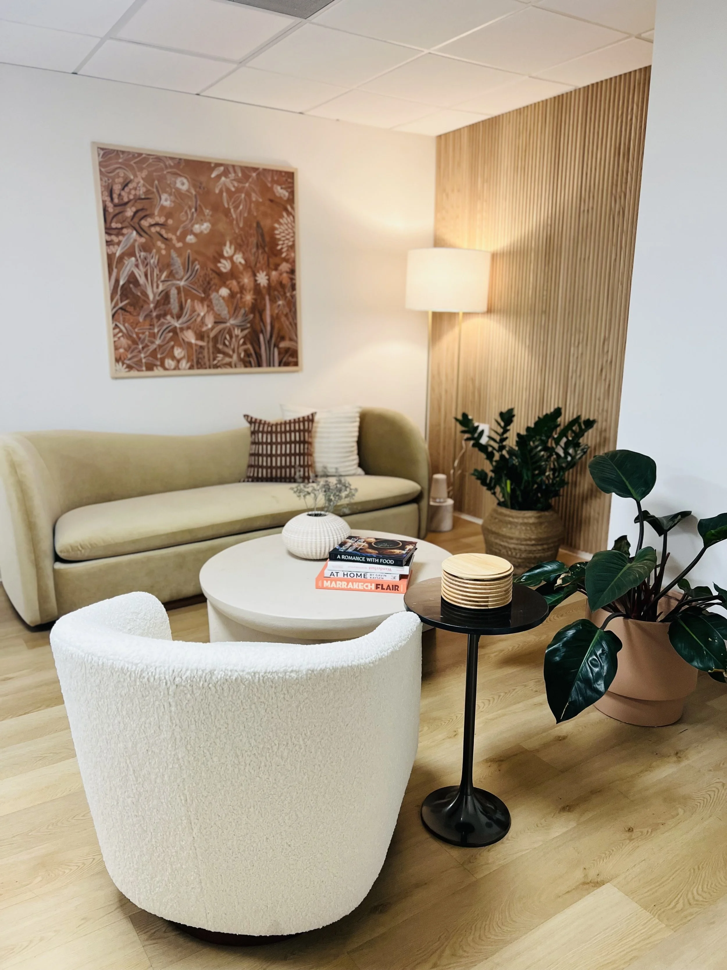 Living room with beige sofa, white armchair, round coffee table with books, large plant in a pink pot, wooden panel accent wall, wall art, and soft lighting.