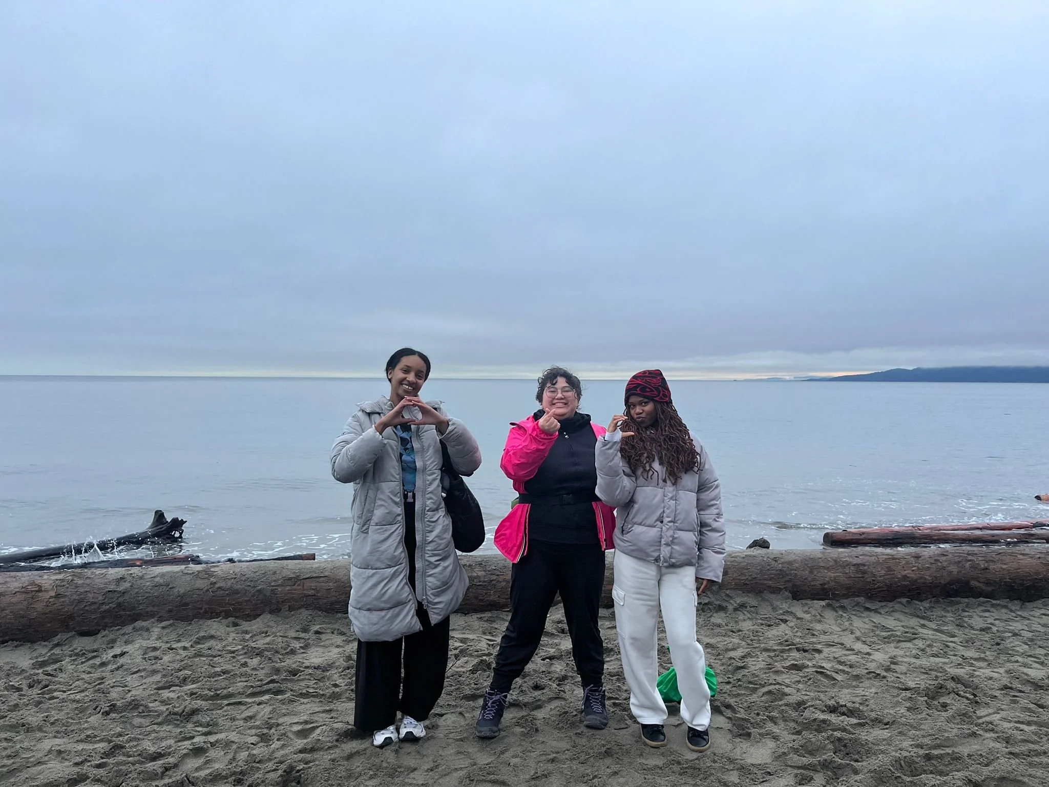 The three women-of-colour founders standing on a sandy beach near the water with a cloudy sky in the background. Each making a letter "O" , "C" and a heart.