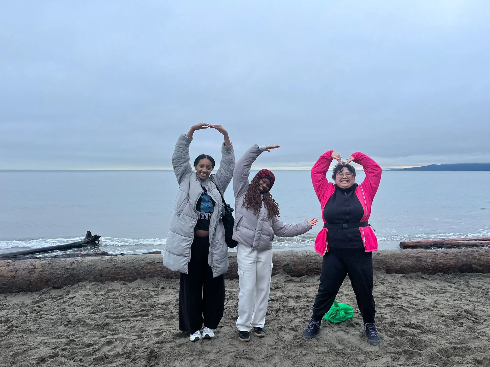 The founders stand on Wreck beach- a sandy beach with logs and ocean behind them.  First is an Ethiopian woman making the letter O. Then, a Kenyan woman making the letter C. Last, a Mexican woman making a heart sign with her arms.