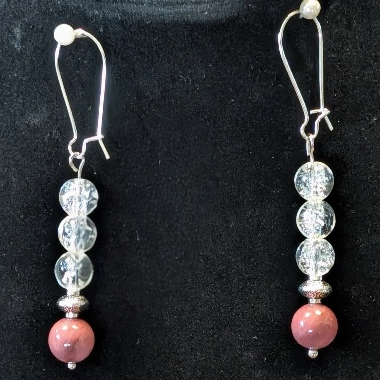 A pair of silver dangling earrings with clear circular beads and pink spherical beads at the bottom, hanging against a black background.
