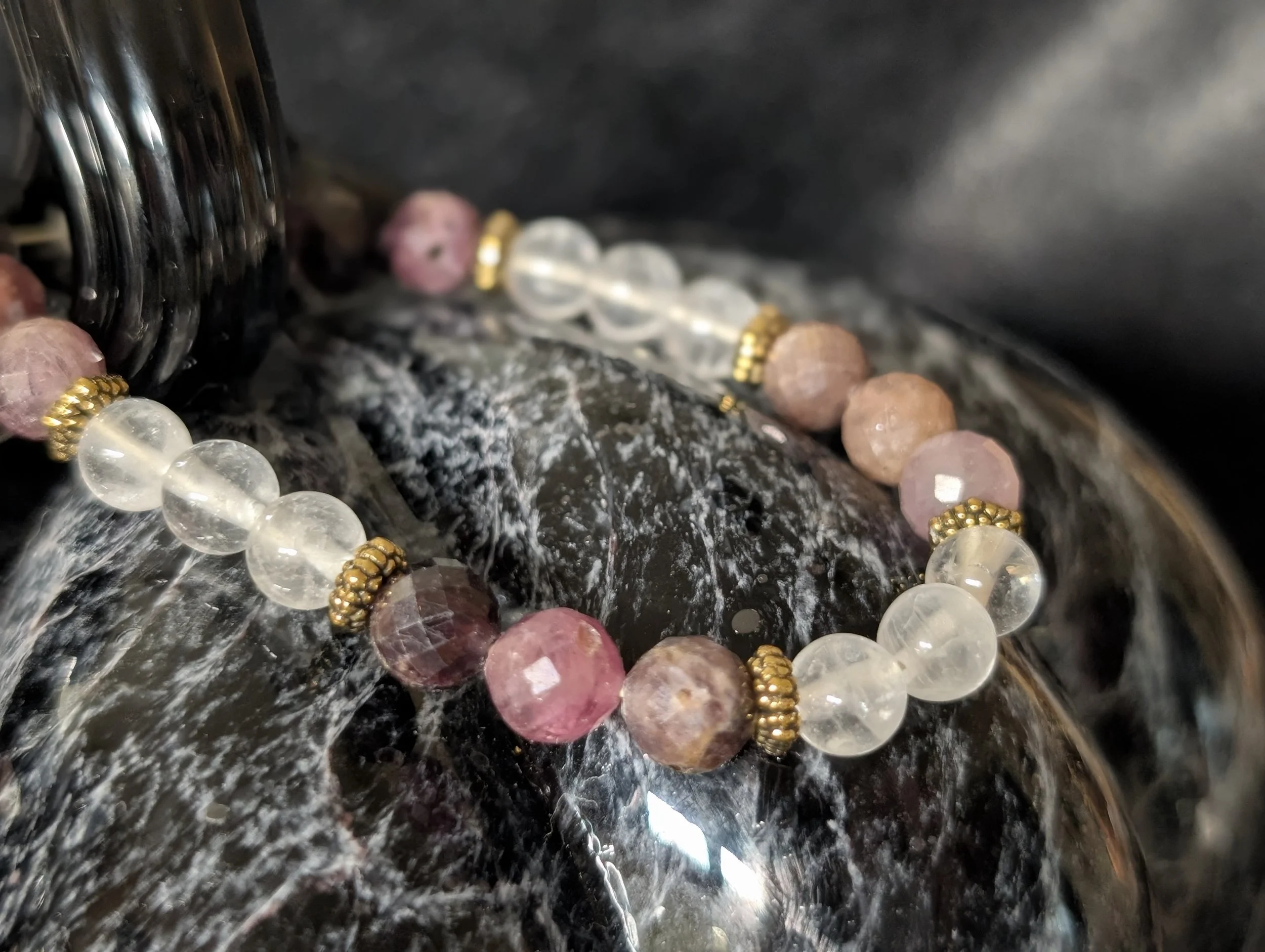 Close-up of a beaded bracelet with pink and clear beads, gold accents, resting on a black textured surface.