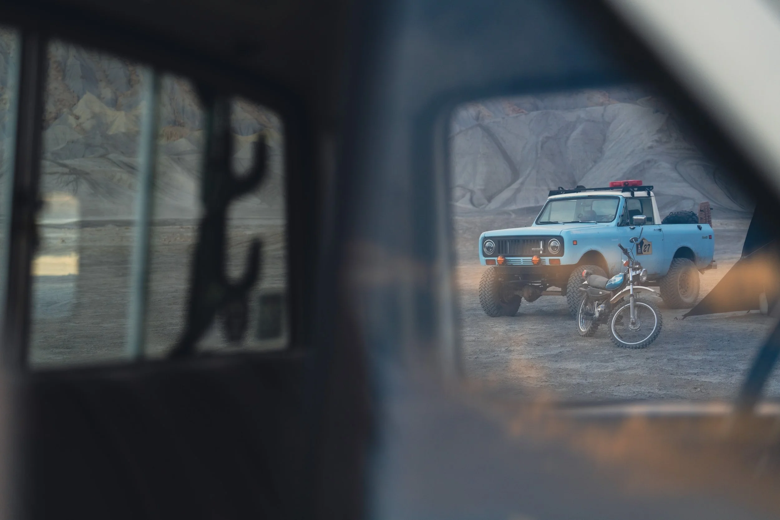 A vintage light blue pickup truck and a black dirt bike parked on a dusty terrain seen through the window of another vehicle.
