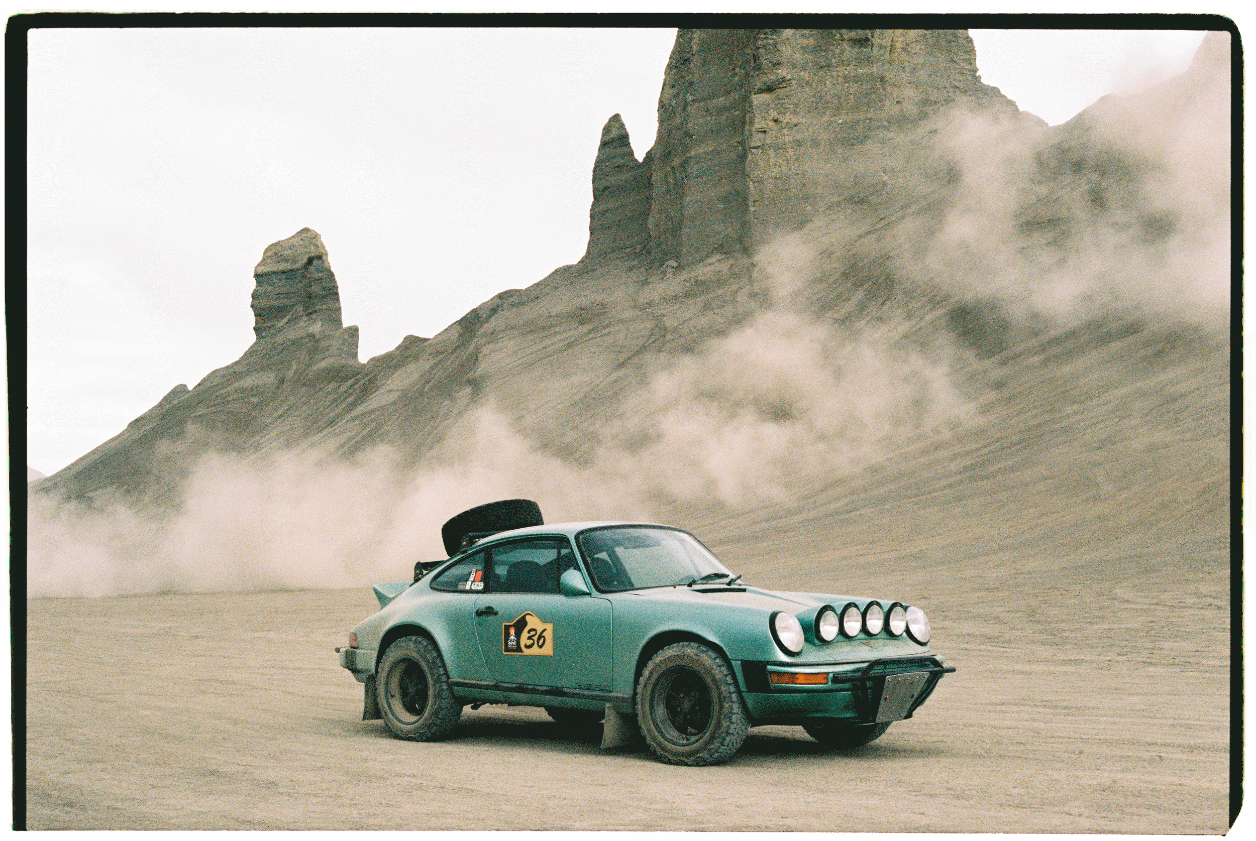 A vintage green Porsche rally car with four front lights and a spare tire on the back, driving on a dusty desert landscape with rock formations and mountains in the background.