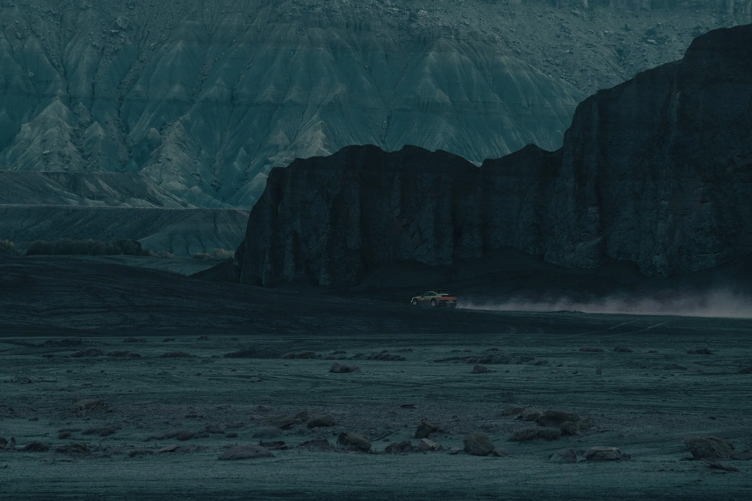 A car driving across a barren, dusty landscape with towering dark rock formations and mountains in the background.
