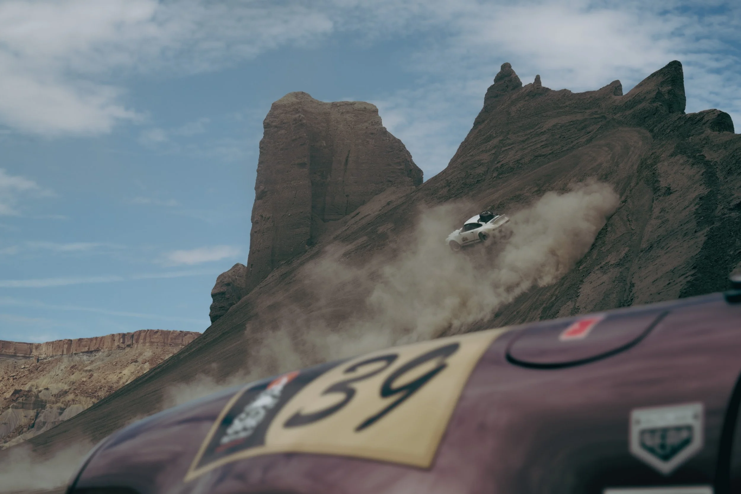 A white car crashing into a rocky hillside in a desert, with a cloud of dust rising around it. In the foreground, part of a racing car with the number 59 is visible.