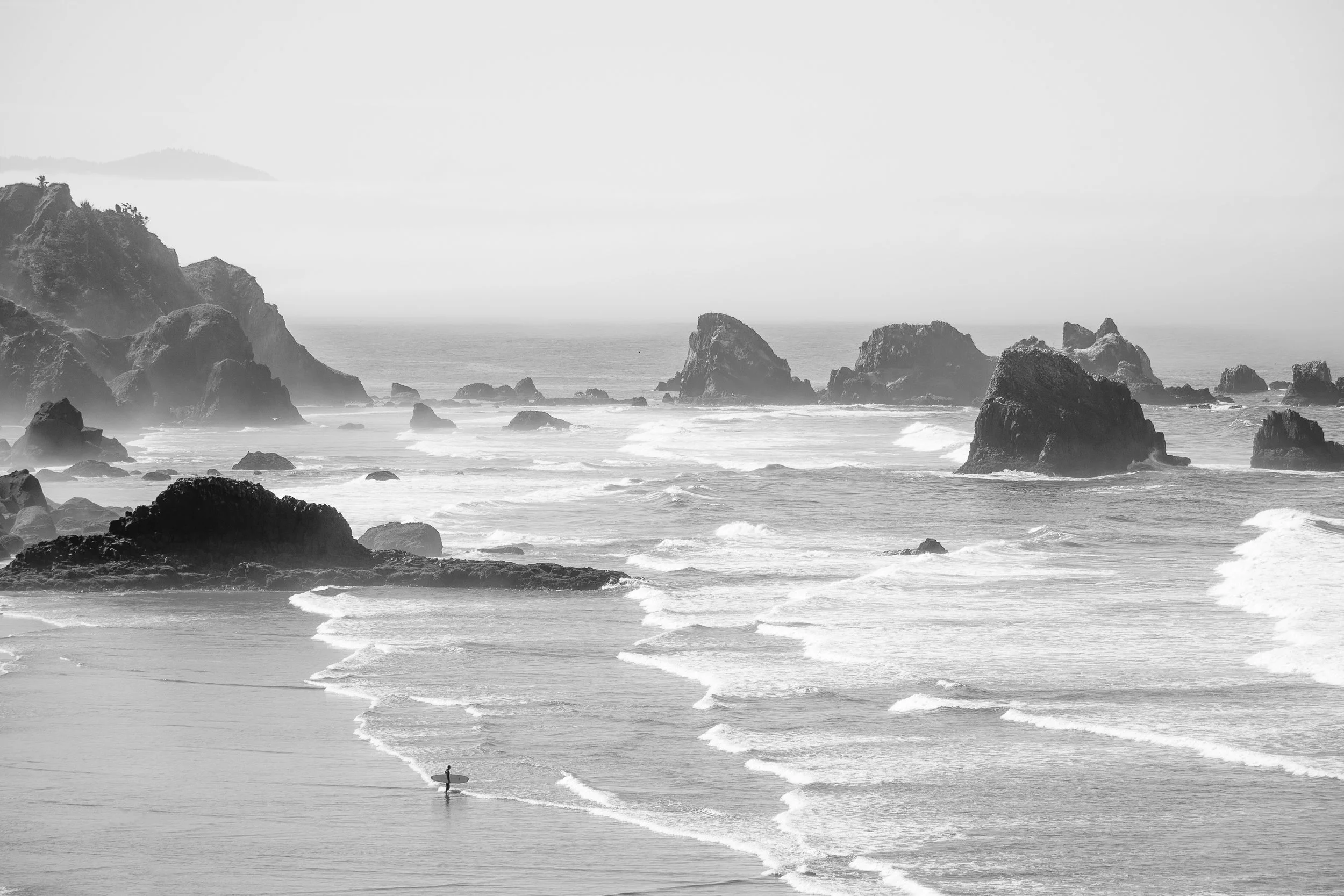 Black and white photo of a surfer walking along the shoreline with waves and rocks in the background.