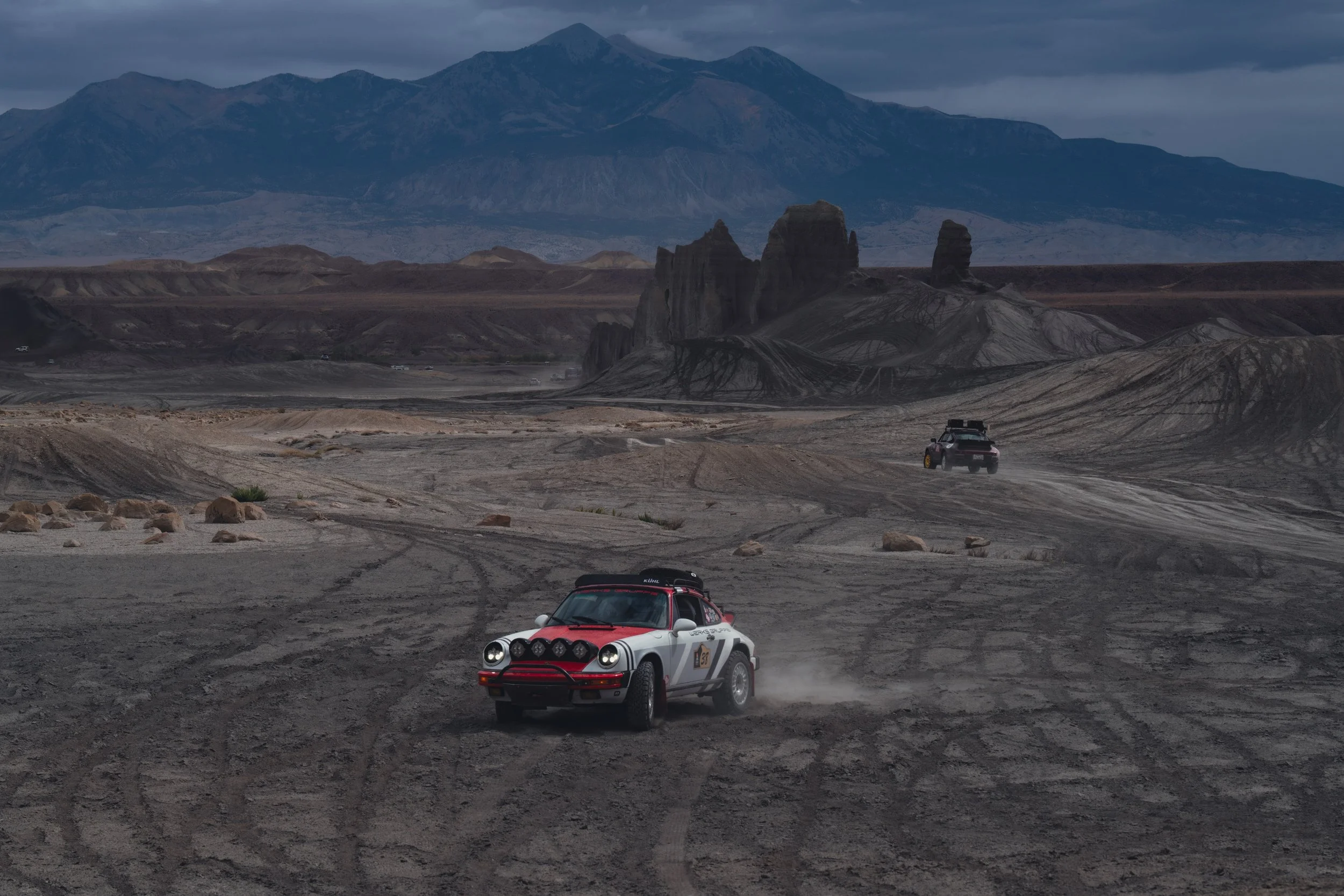 Two off-road rally cars driving through a dusty desert landscape with mountains and rock formations in the background under a cloudy sky.