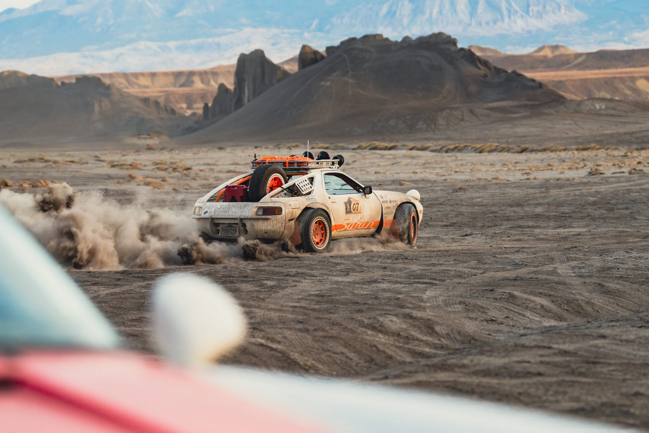 A rally or race car kicking up dust in a barren desert landscape with rocky mountains in the background.