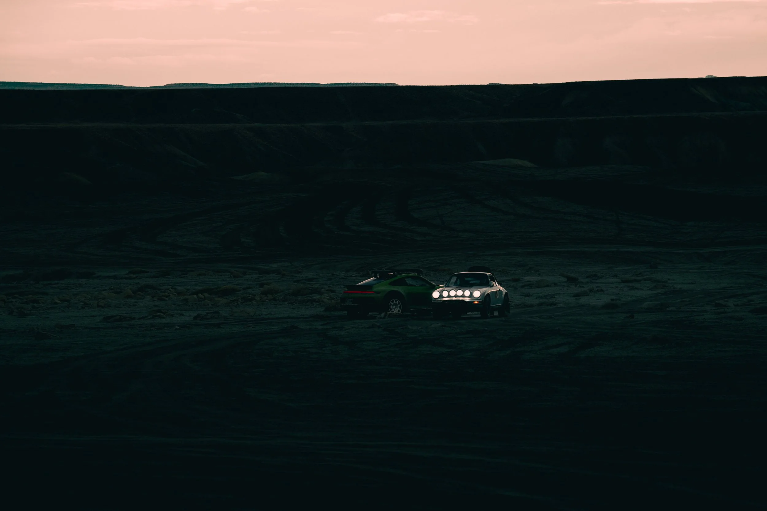 Two cars parked on a dark, barren landscape with tire tracks, silhouette of a hill in the background, during twilight