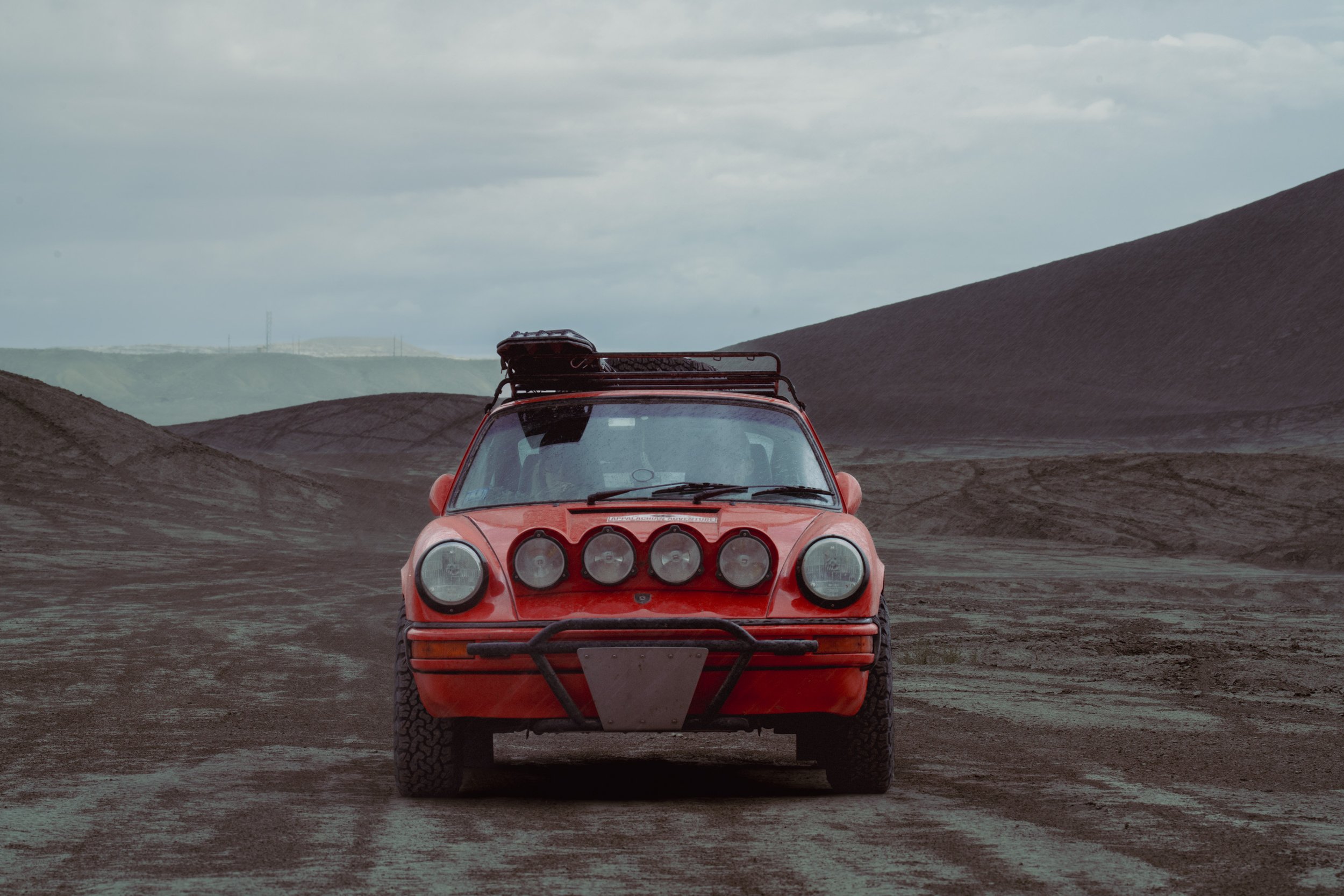 A red vintage off-road vehicle with round headlights and additional front lights driving on a dirt road in a barren, mountainous landscape under a cloudy sky.