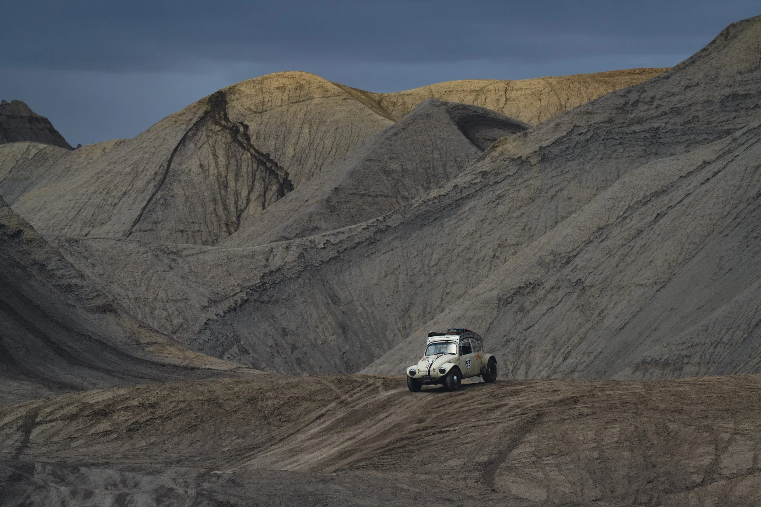 A vintage white car parked on a dirt road surrounded by barren, rugged mountains with dark clouds overhead.
