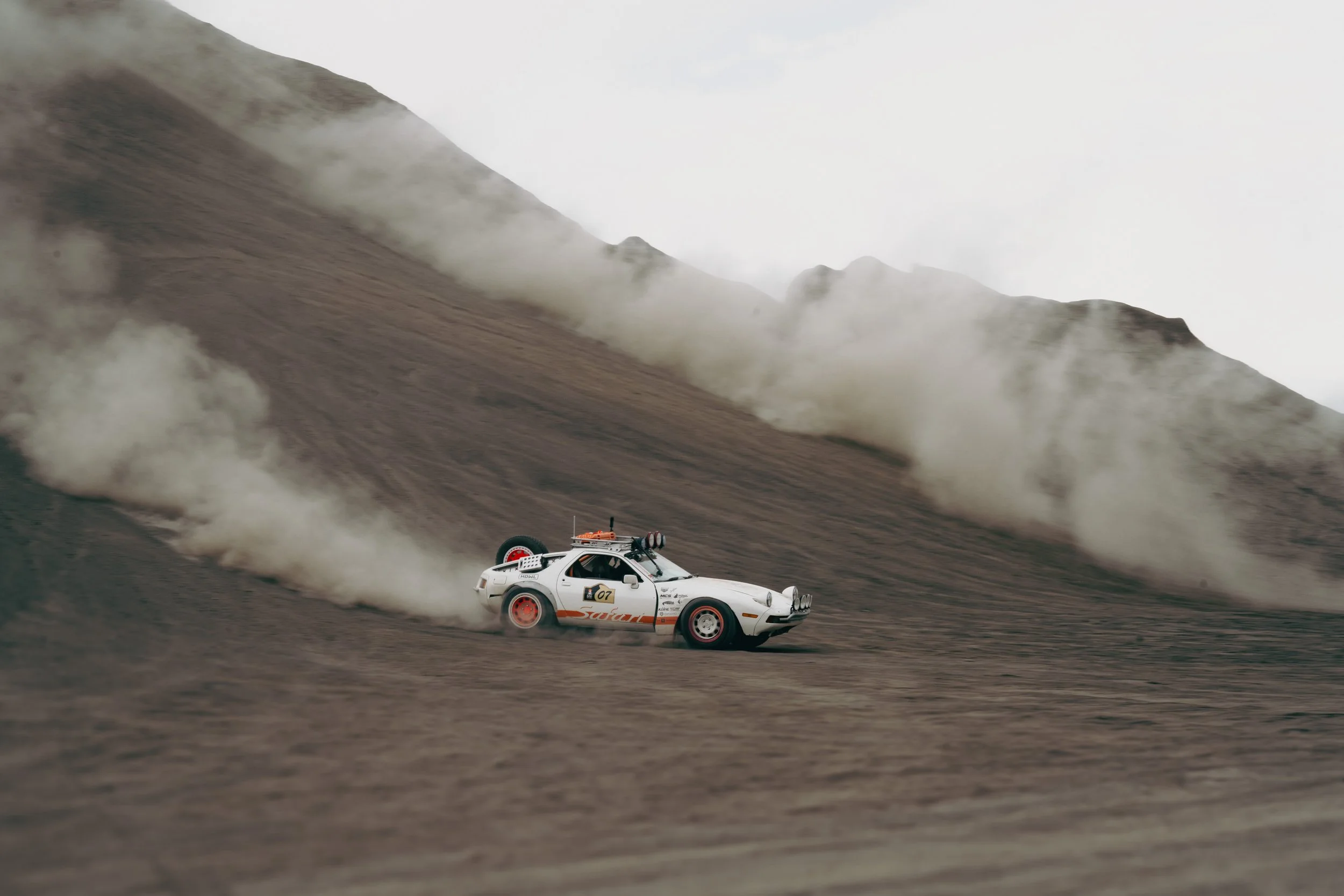 A white rally car driving on a dirt track with dust clouds behind it, mountainous landscape in the background.