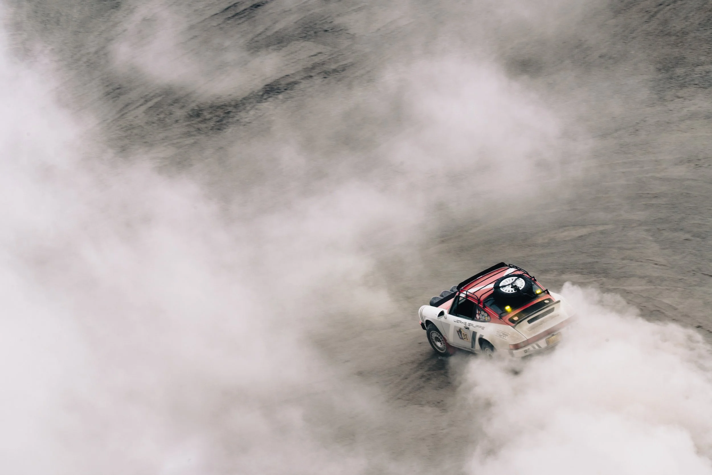 A rally car driving through a dusty dirt track, kicking up a large cloud of dust behind it.