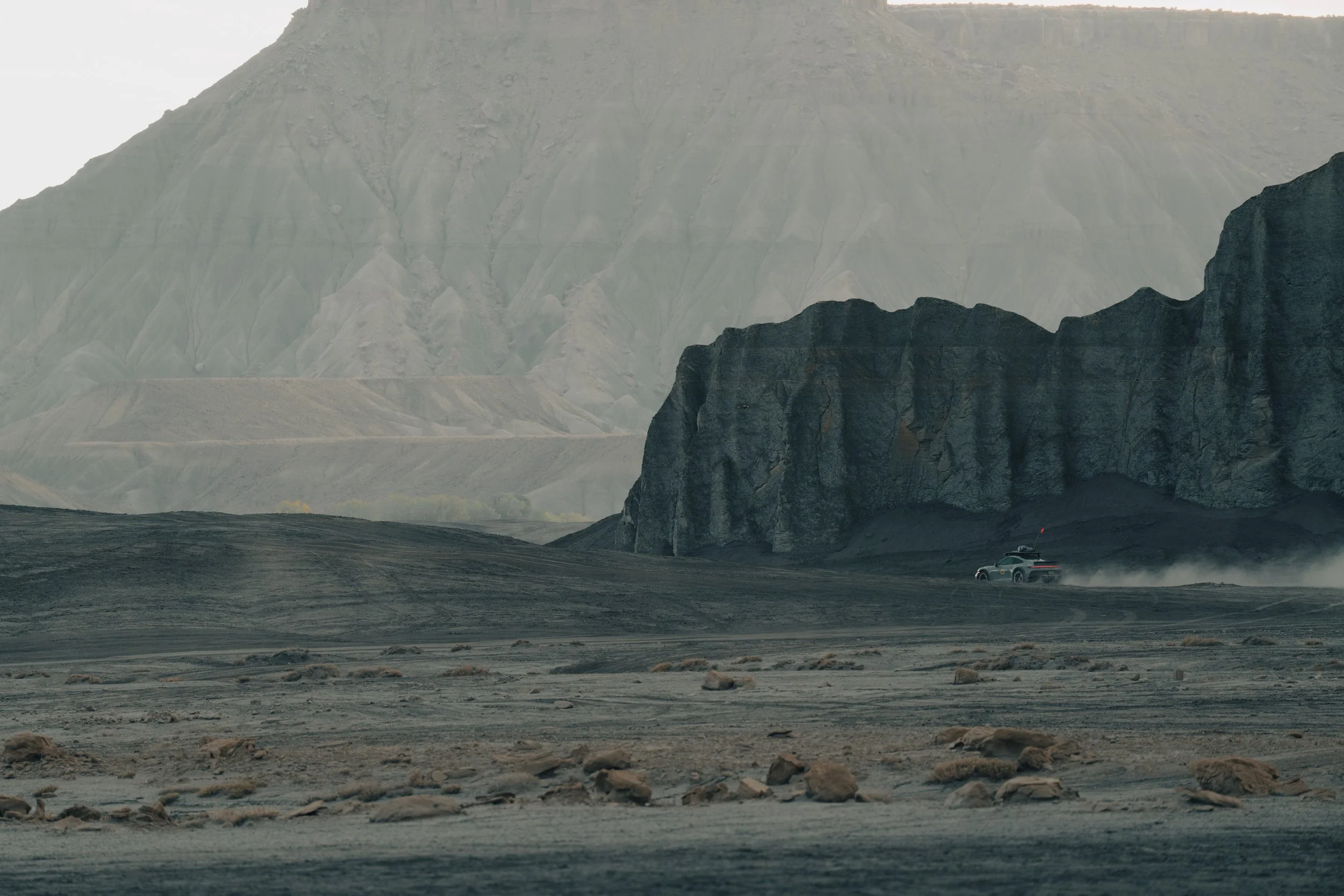A rugged desert landscape with dark, rocky formations and mountains in the background. A vehicle is driving across the terrain, kicking up dust.