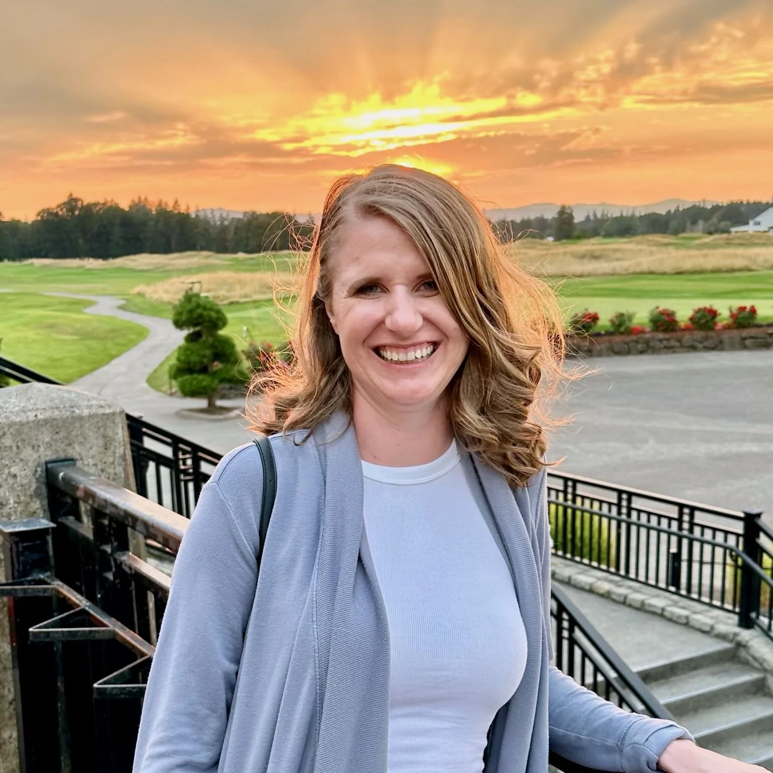 A woman smiling outdoors during sunset, standing on a staircase with a garden and open field in the background.