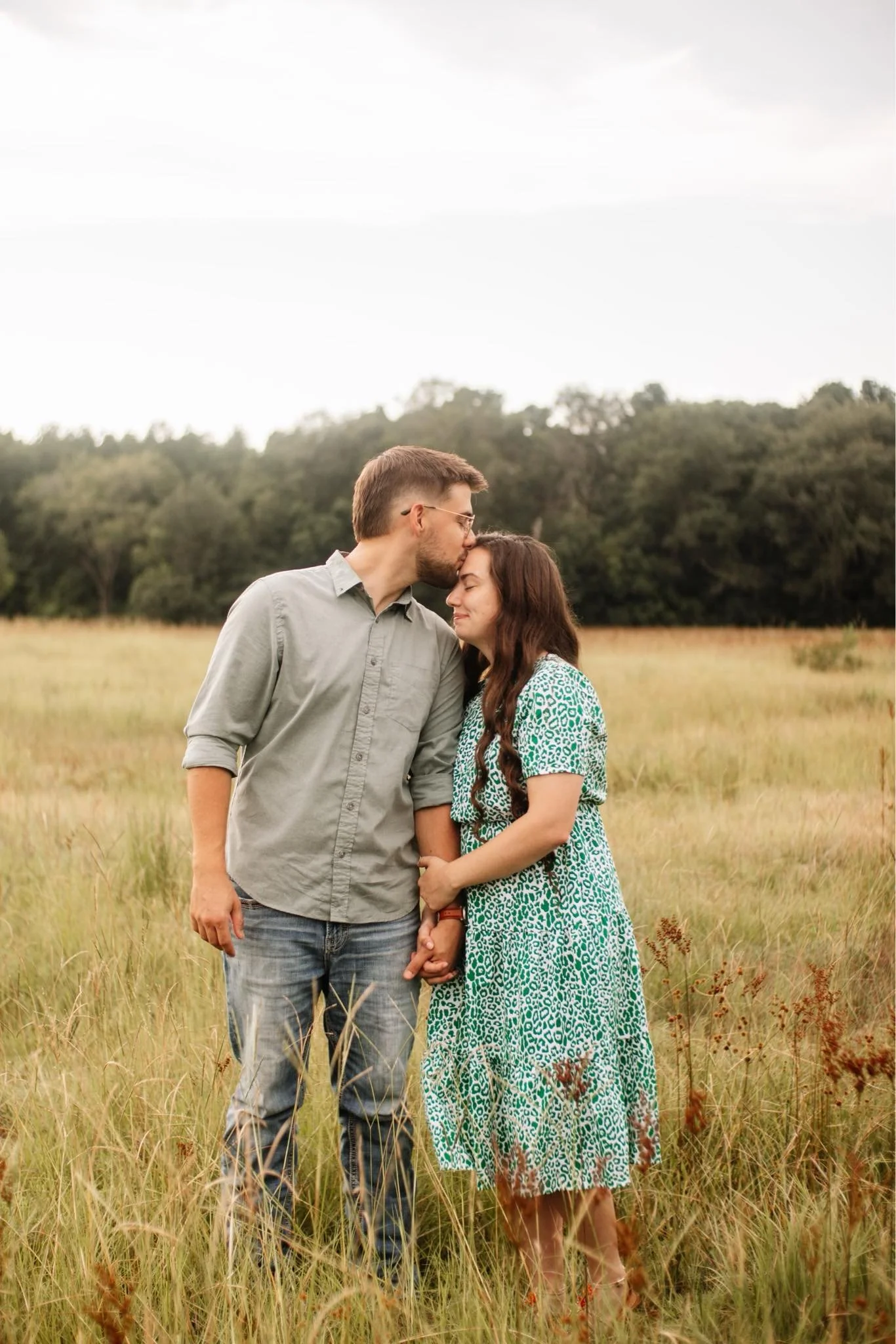 A couple standing in a grassy field, the man kissing the woman's forehead as they hold hands and smile with trees in the background.
