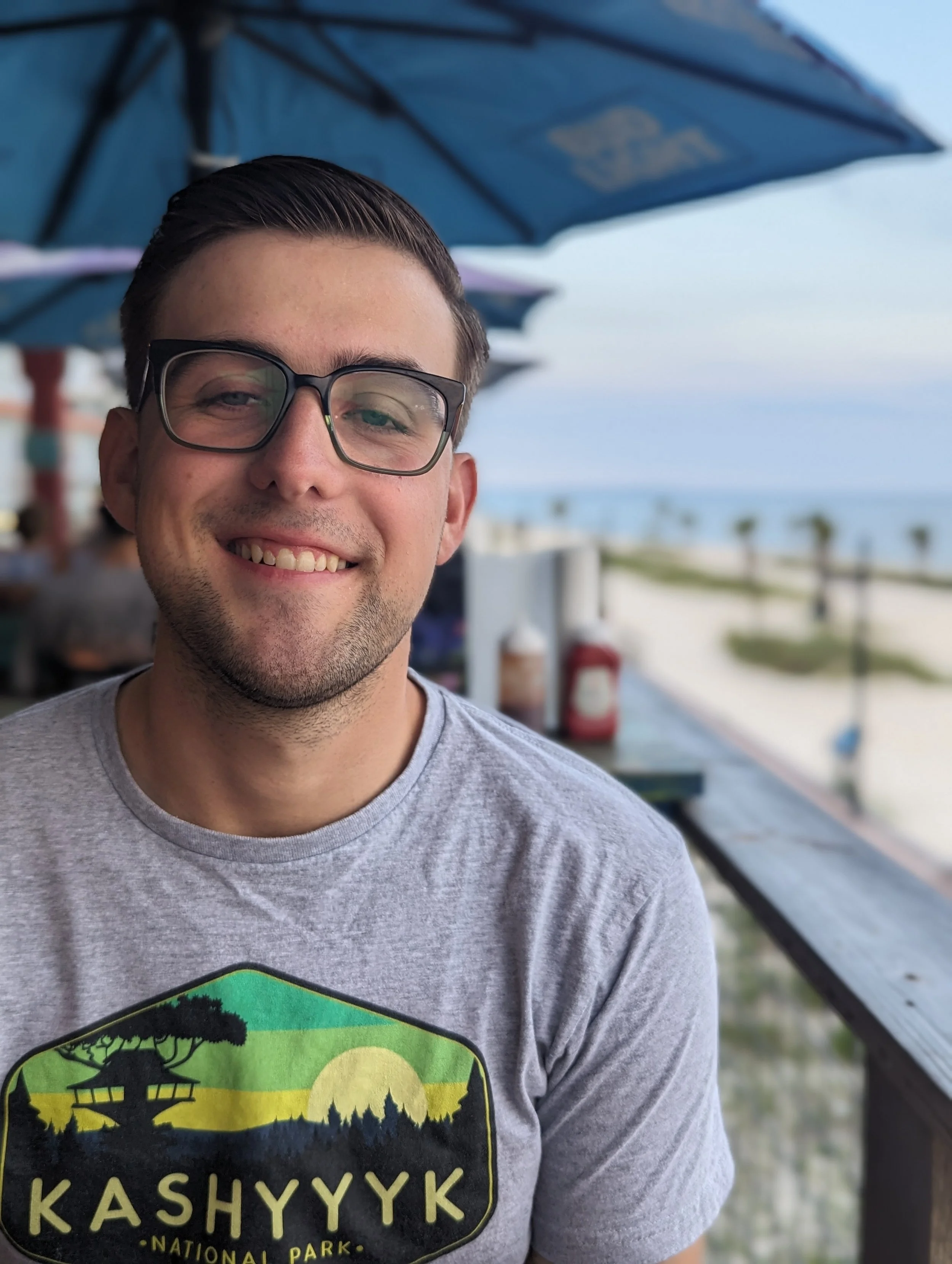 A smiling man with glasses and a gray T-shirt featuring a colorful KASHYYYK National Park logo, sitting outdoors near a beach, under a blue umbrella.