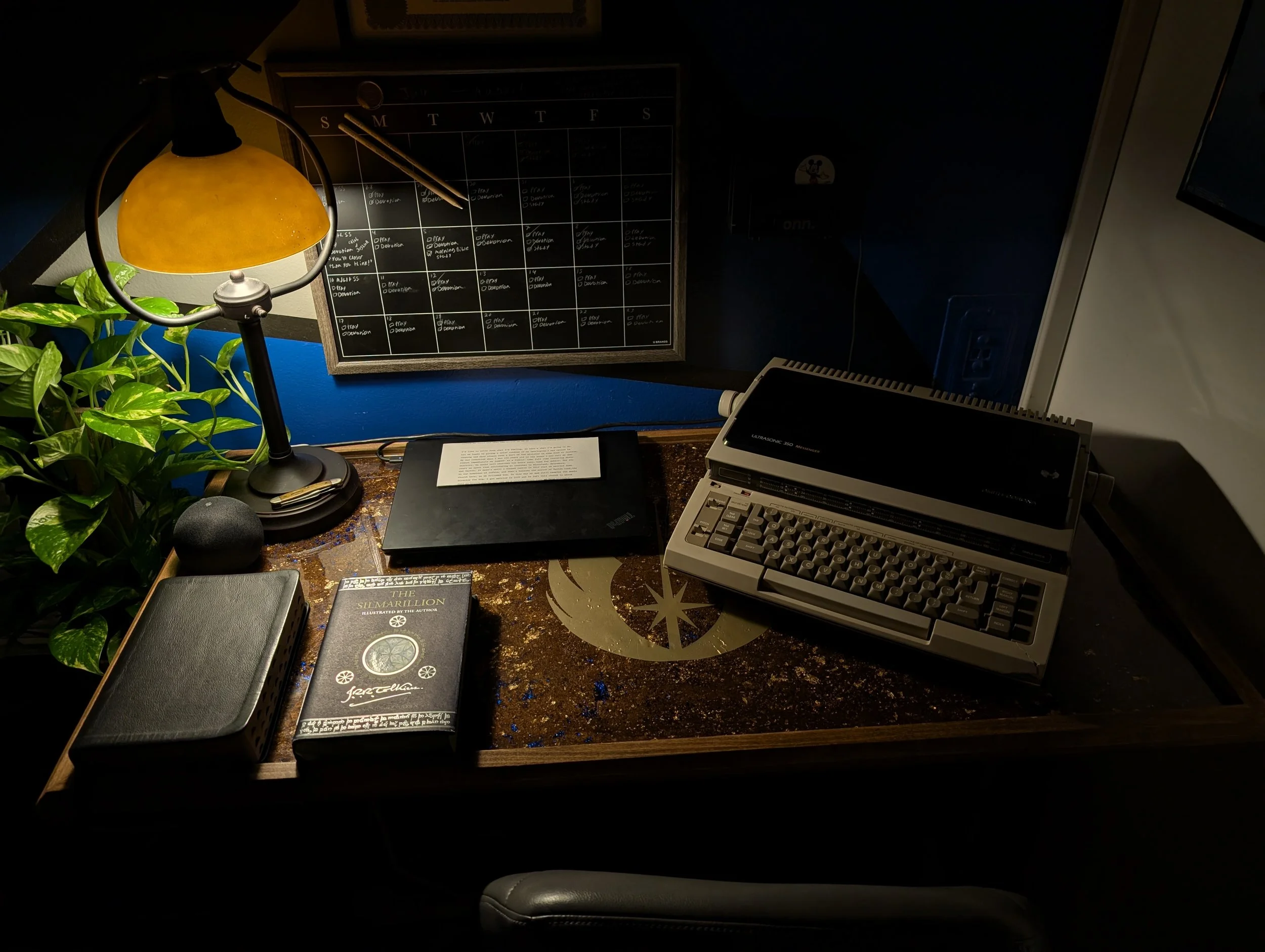 An organized workspace featuring a desk lamp, a potted plant, a closed black notebook, a book titled "The Silmarillion," an electronic device, and a vintage typewriter on a desk with a decorative star and moon design. A wall calendar and power outlet are visible in the background.