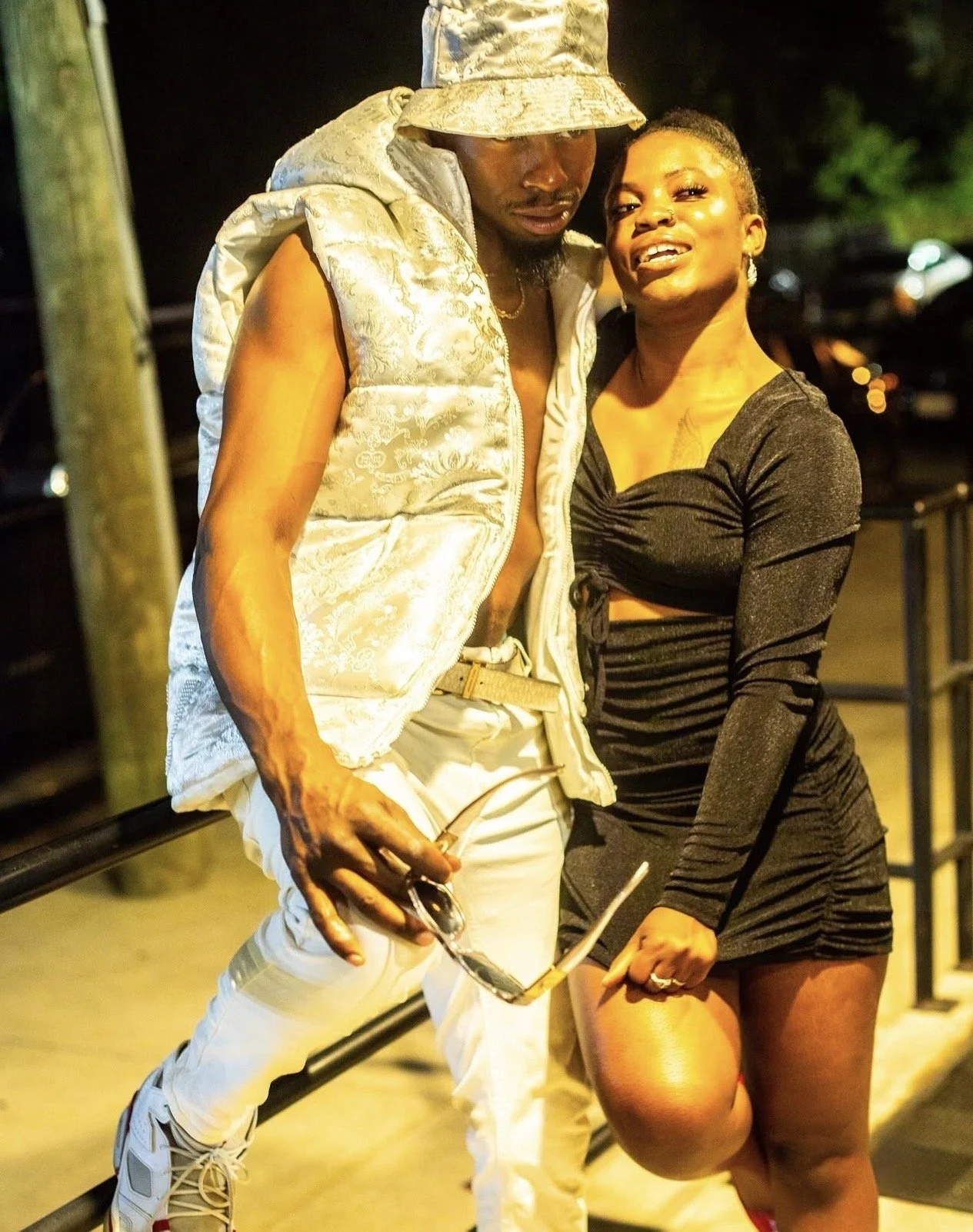 A man and woman standing outdoors at night, posing together. The man is wearing a beige hat, a puffy vest, and light-colored pants. The woman is wearing a fitted black dress with long sleeves and is smiling at the camera.