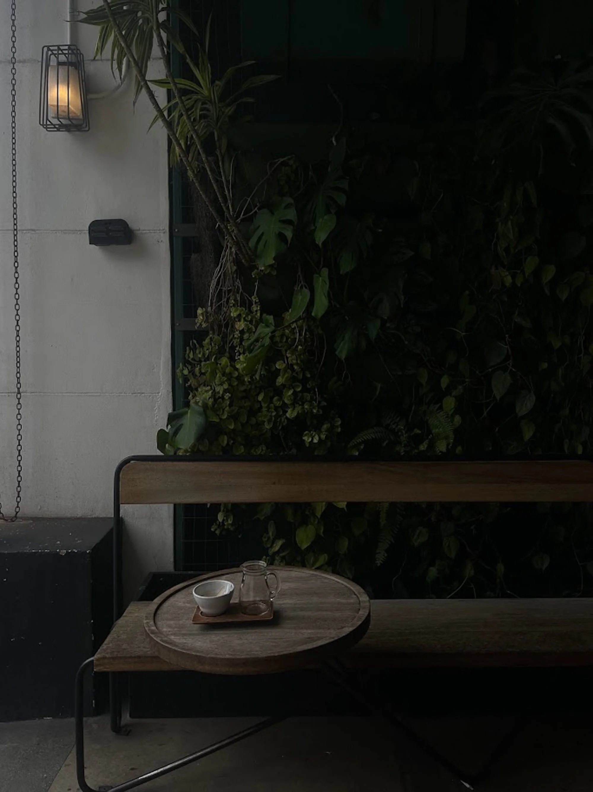 Outdoor patio scene with a wooden table set for coffee, surrounded by lush green plants and a white wall with a wall-mounted lantern and a black device, under warm lighting.