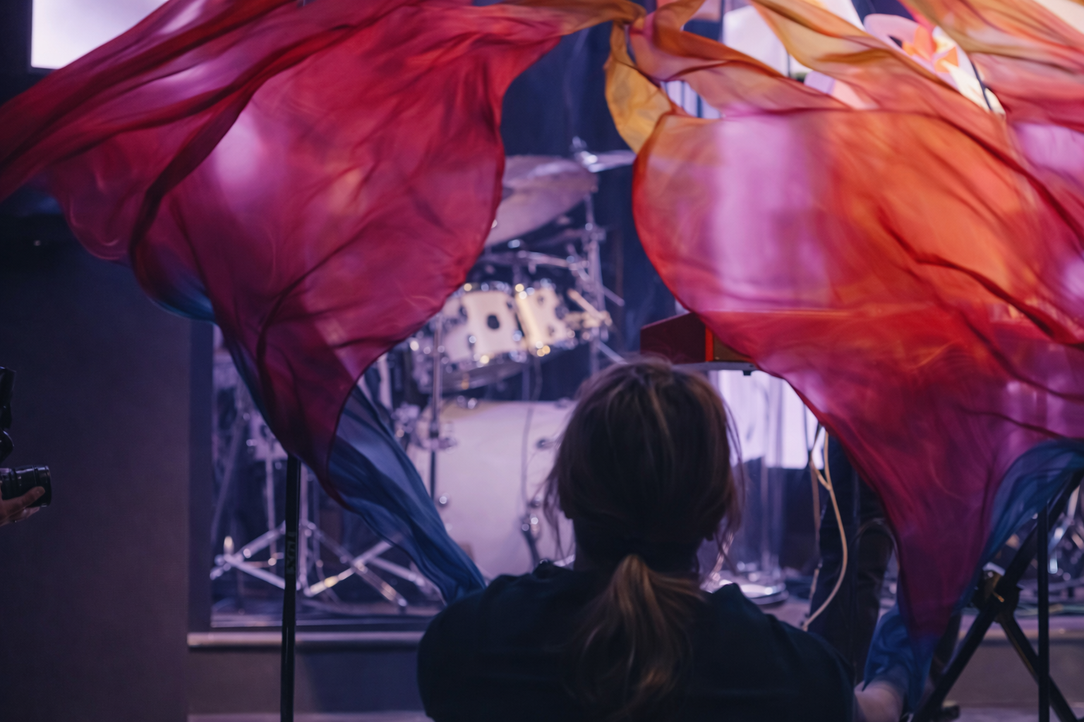 Person sitting in front of a stage with large colorful fabric decorations hanging down, behind a drum set and music equipment.