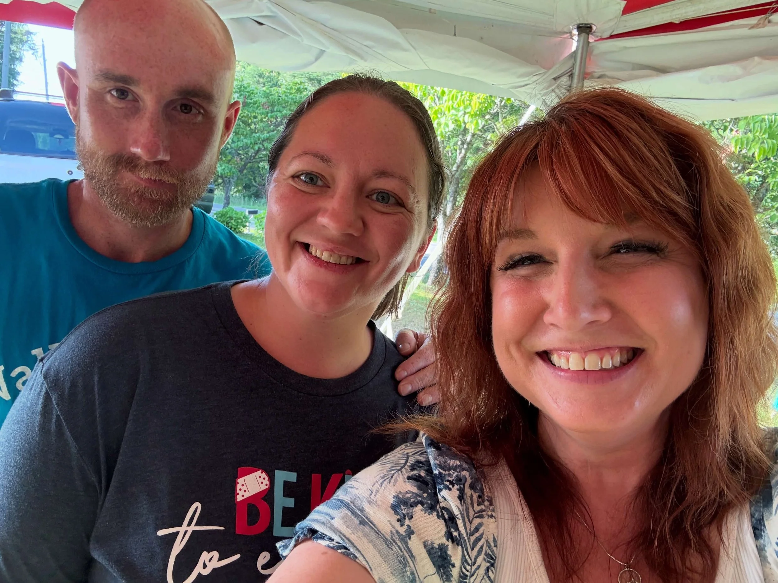 Three people taking a selfie outdoors under a canopy, with trees and a bus in the background, smiling and happy.